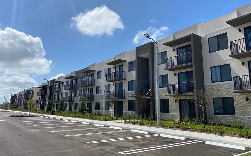 Modern apartment building with balconies and a parking lot in front, under a blue sky with clouds.
