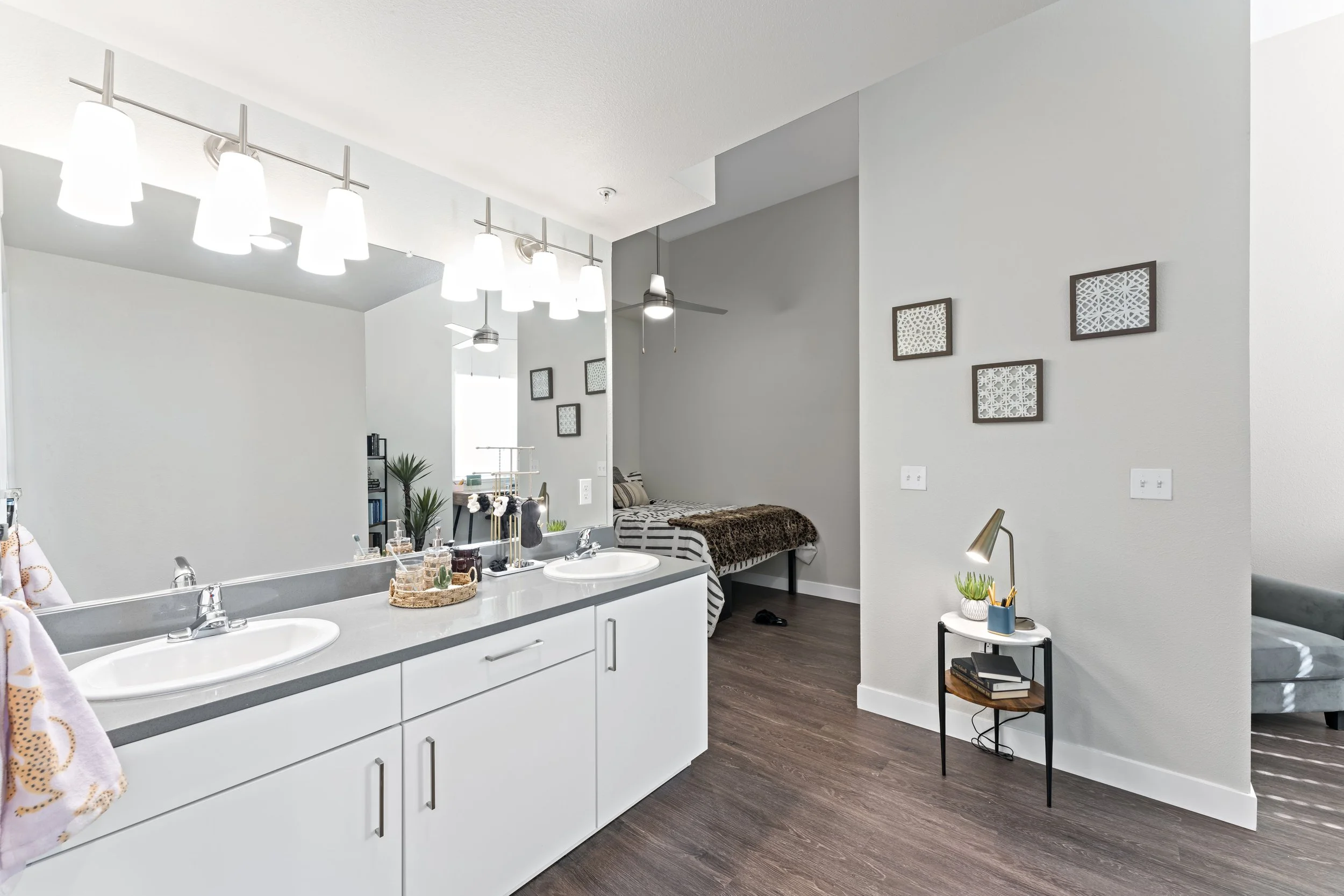 Modern bathroom with double sink vanity, white cabinets, large mirror, decorative items, and bedroom with bed and wall art in background.