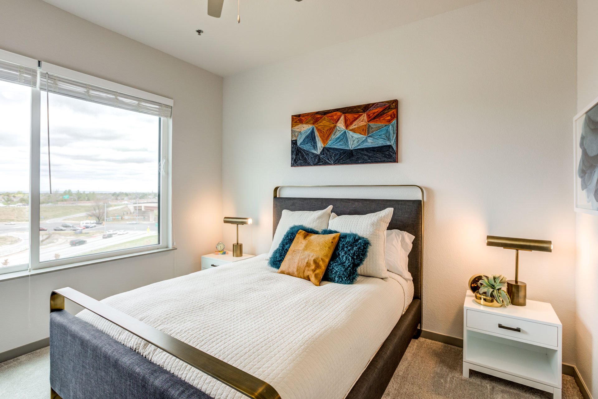 Bedroom with a large window, white walls, and a bed with a gray headboard. The bed has white bedding and decorative pillows, including a blue furry pillow and a tan pillow. There are two white nightstands with gold lamps, a small clock, and a plant. 