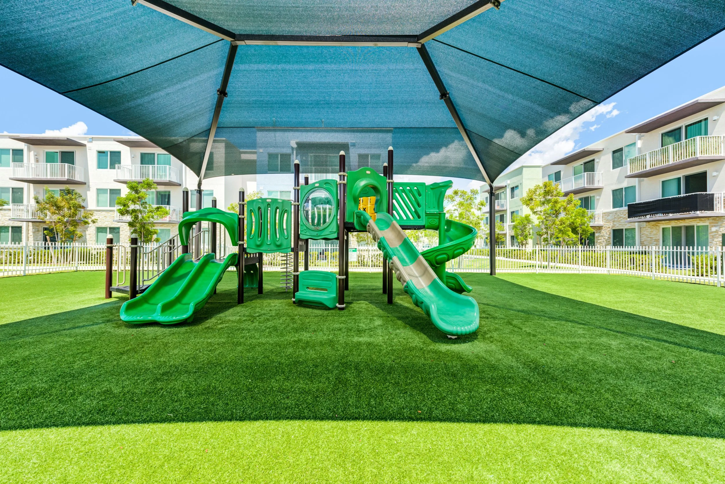 Children's outdoor playground with green slides and climbing structures, shaded by a large tent, surrounded by a grassy area and apartment buildings.