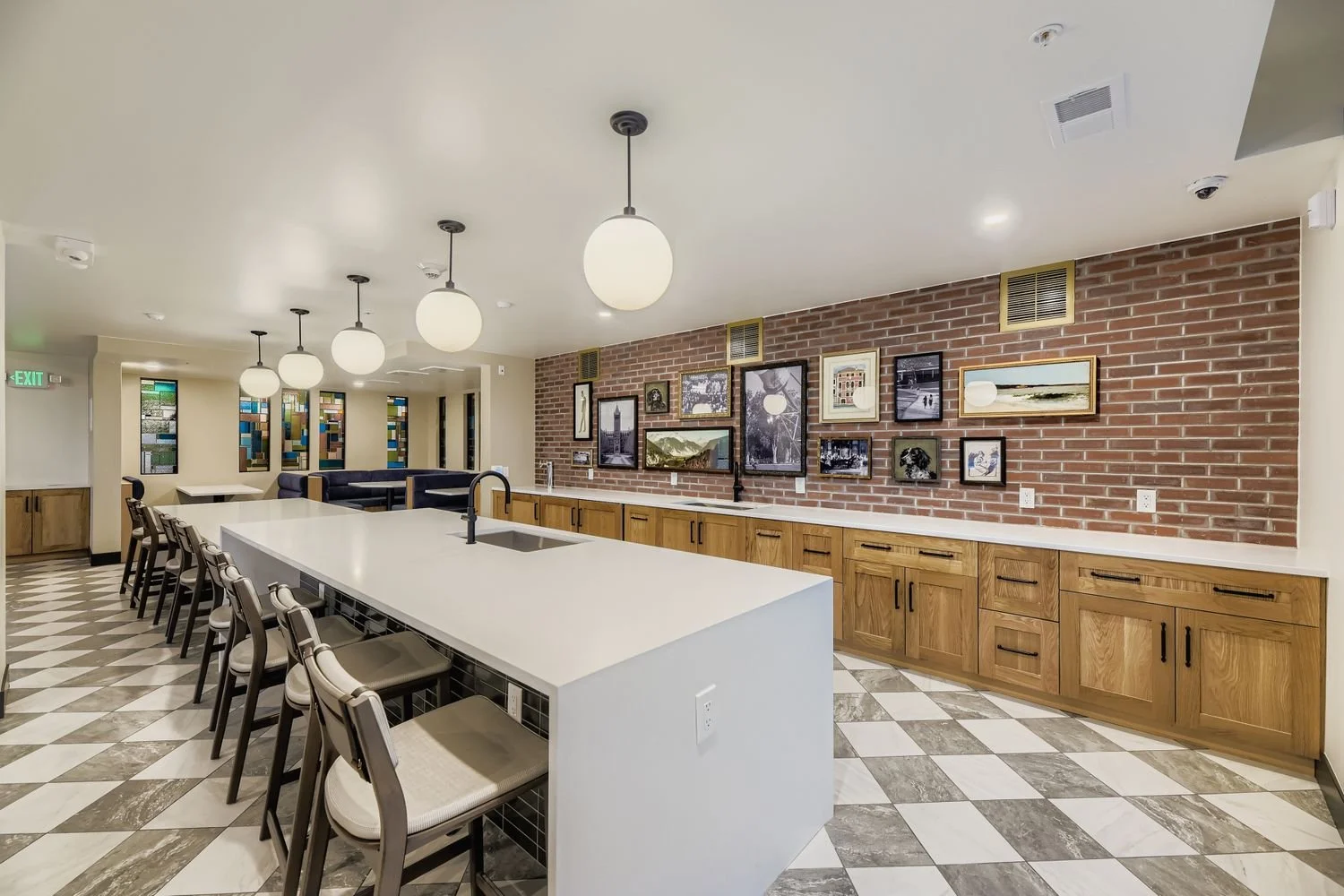 Modern kitchen with a large white island, wooden cabinets, brick wall with framed artwork, tiled floor, and white ceiling with hanging spherical light fixtures.