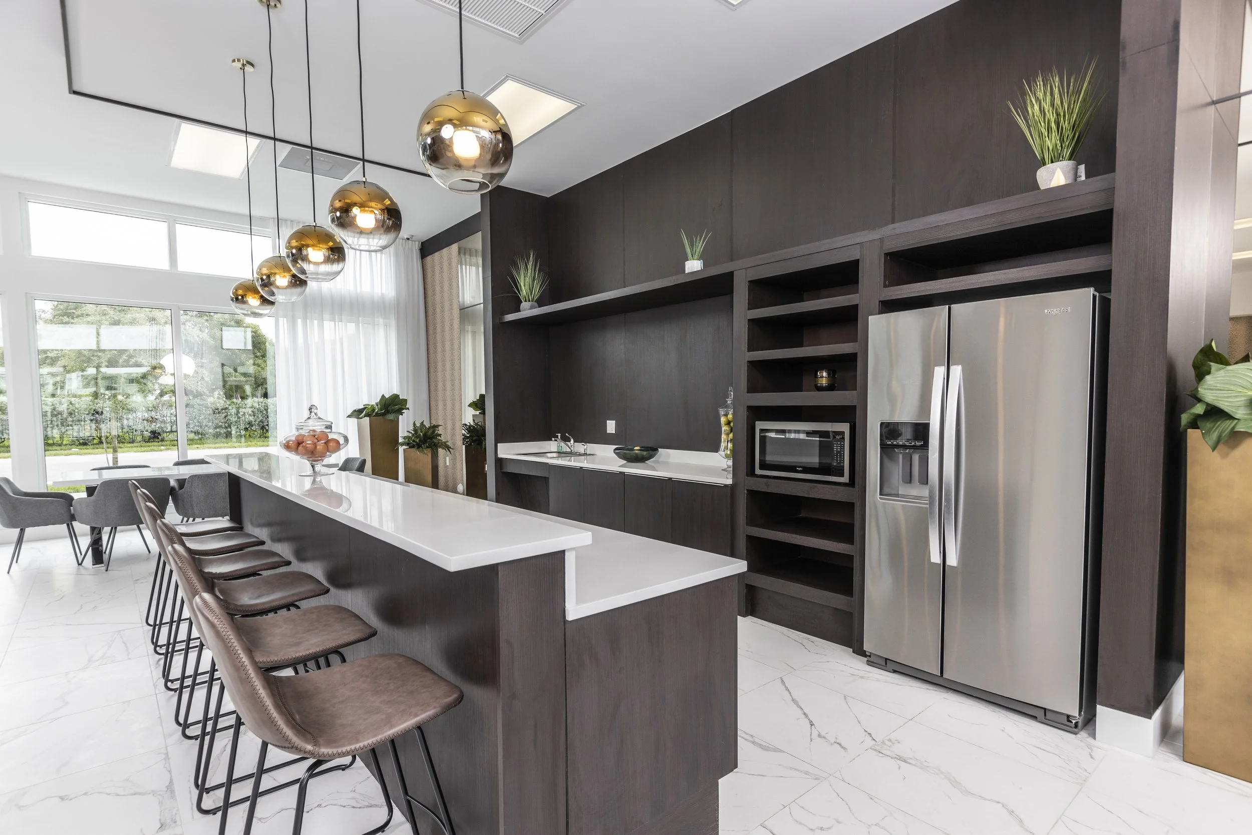 Modern kitchen with a white island, dark wood cabinets, stainless steel refrigerator, microwave, and pendant lights, with large windows and green plants.