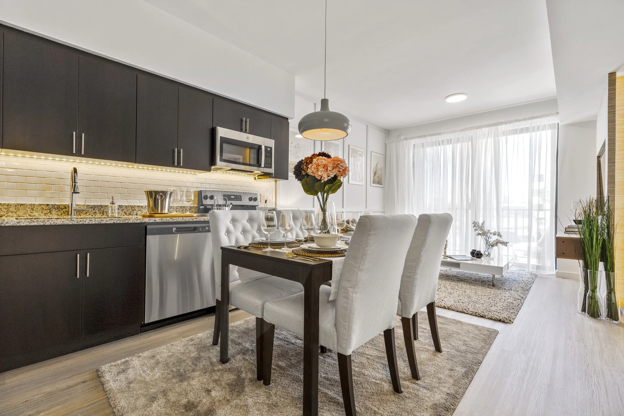 Modern open-concept kitchen and dining area with white walls, dark cabinets, granite countertops, and a dining table set with white upholstered chairs, wine glasses, and a vase of pink and orange flowers.