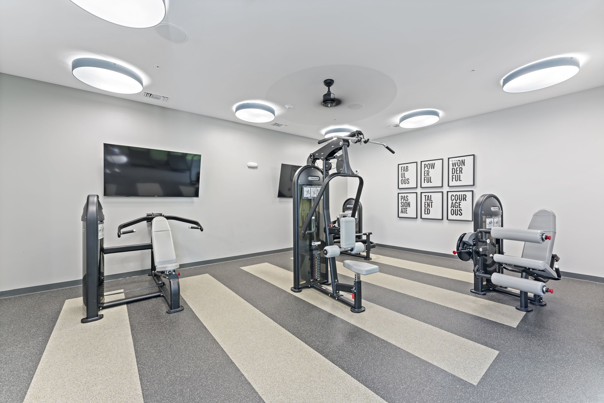 Fitness room with weight machines, flat-panel TV on wall, motivational posters, and ceiling lights, with striped patterned floor.