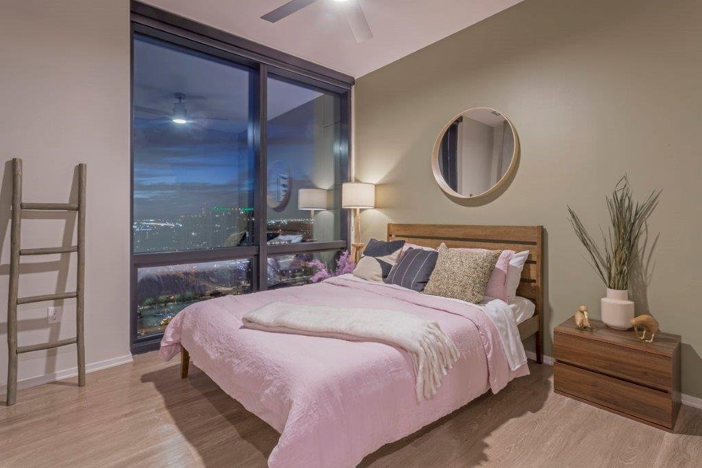 Modern bedroom with large window overlooking city skyline at night, wooden bed with pink bedding, nightstand with decorative items, and a mirror on the wall.