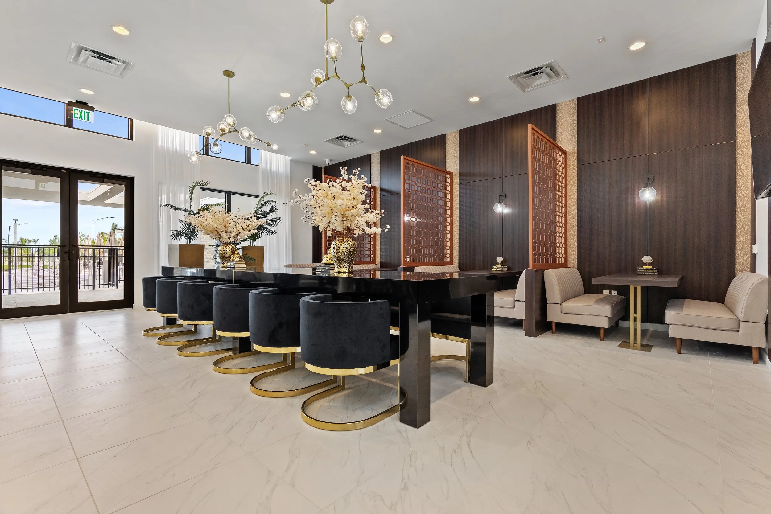 Elegant modern dining area with a long black table, black velvet chairs with gold bases, decorated with white floral arrangements in gold vases. Beige lounge chairs with side tables are along a dark wood wall, which has ambient lighting fixtures. Lar