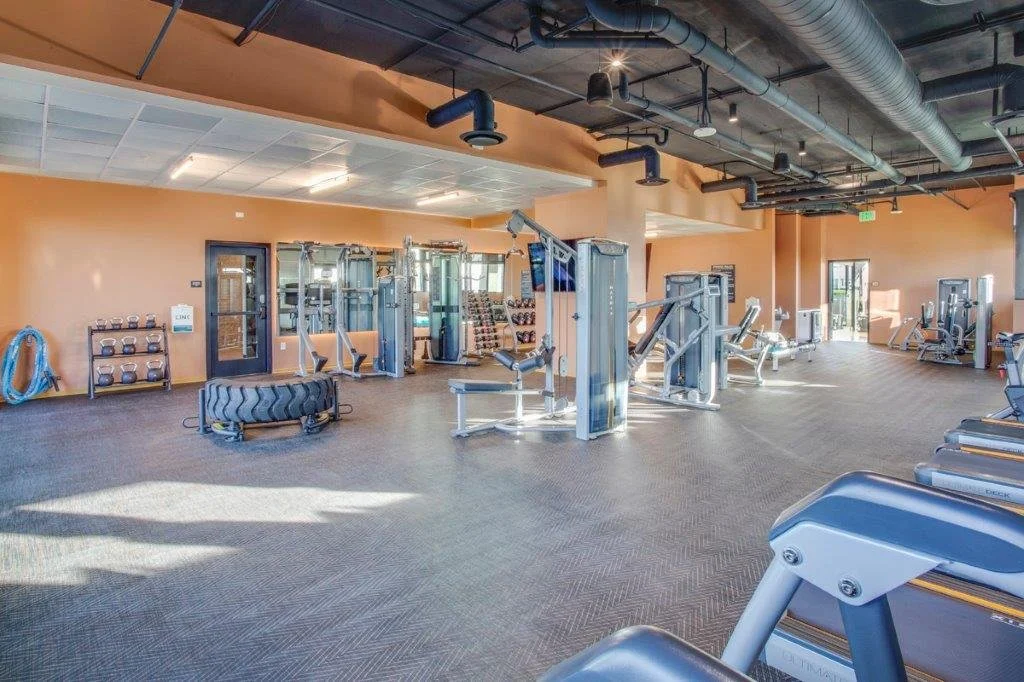 Empty gym with various workout equipment including weight machines, additional cardio machines, dumbbells, and wall-mounted storage for kettlebells and hoses, in a spacious area with a high ceiling and exposed ductwork.