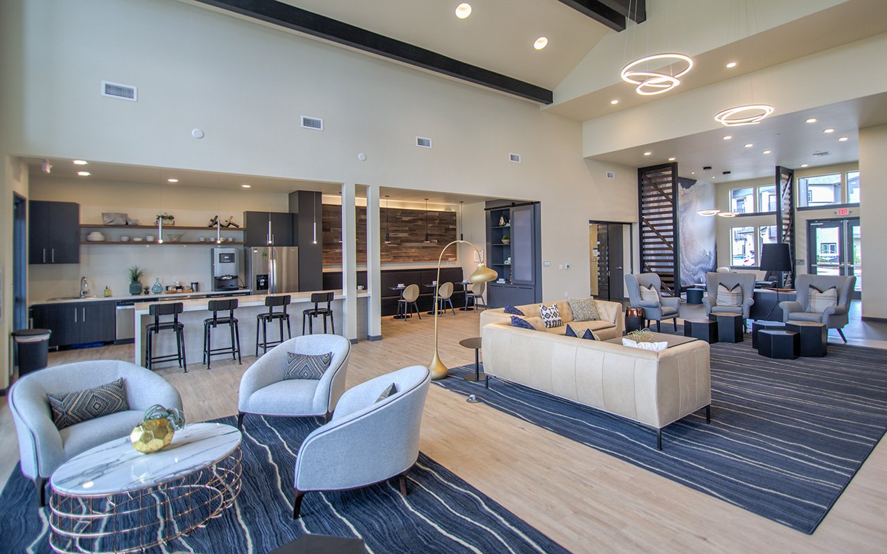 Modern hotel lobby with seating area, bar stools, and open kitchen in the background, high ceilings with circular pendant lights, large windows letting in natural light, and contemporary decor.