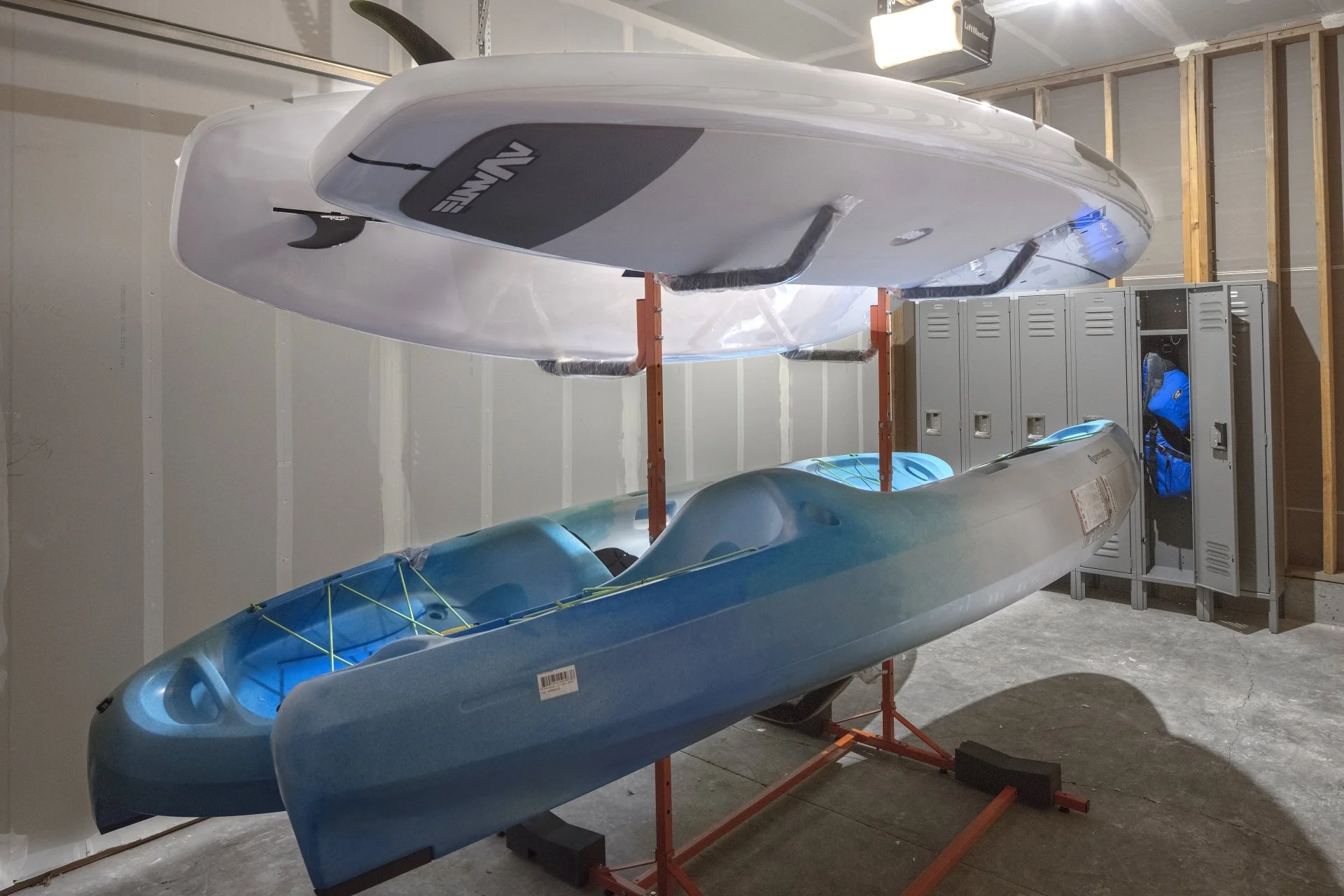 Two blue and white kayaks stored on orange kayak stands, with metal lockers and unfinished wooden wall in background.