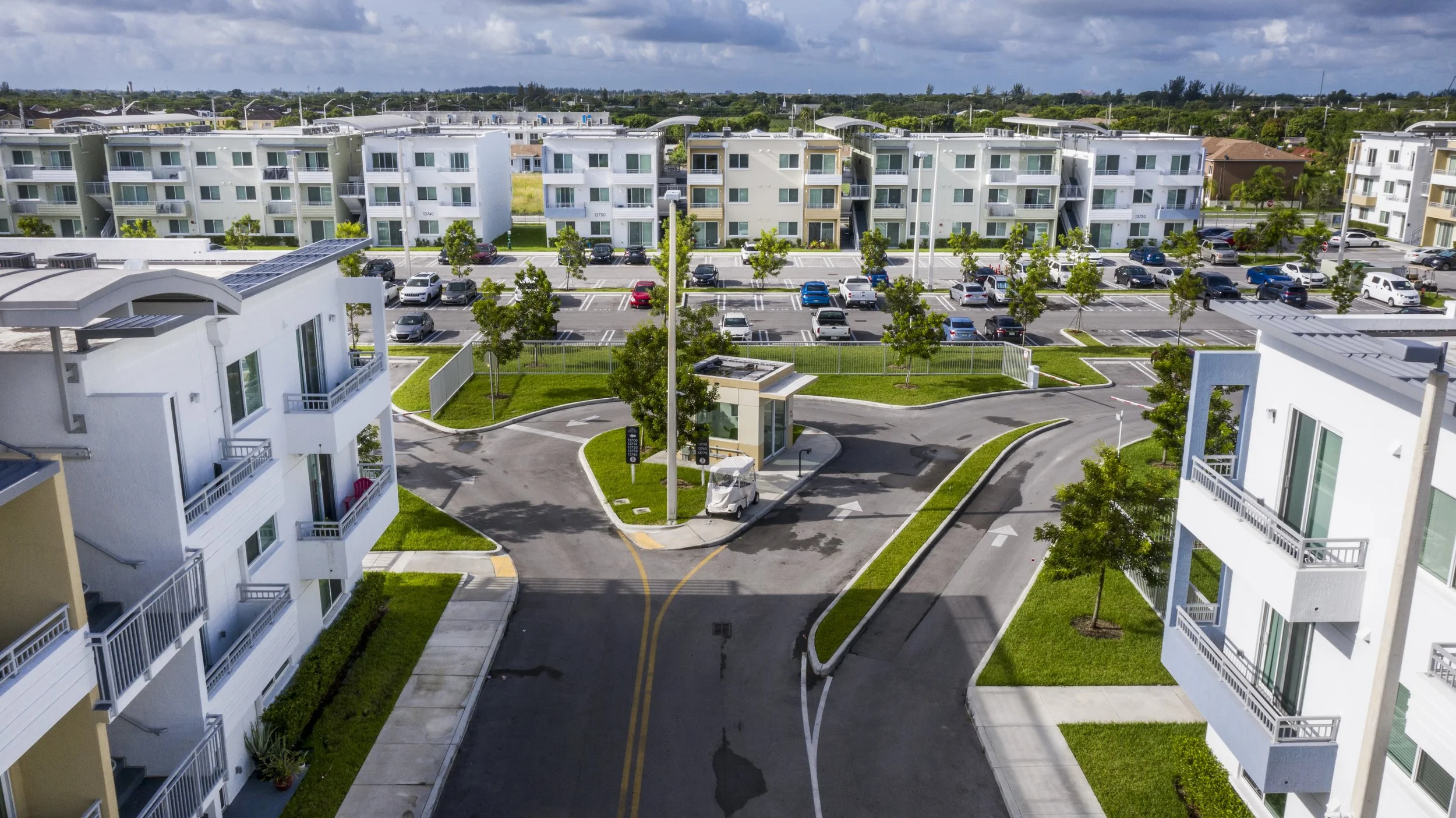 Aerial view of a modern apartment complex with multiple white buildings, green lawns, and parking lots filled with cars.