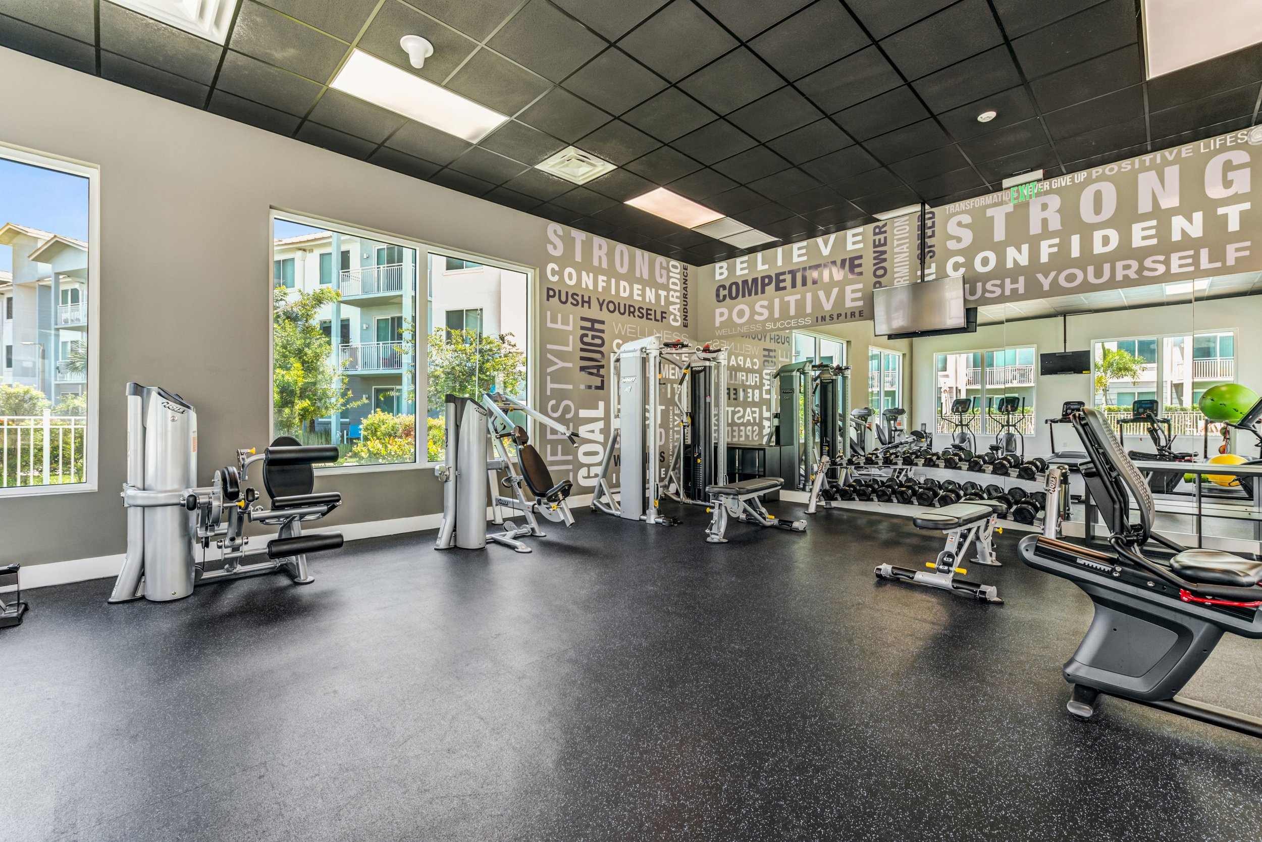 A well-lit gym with large windows, exercise equipment including weight machines, dumbbells, and cardio machines, and motivational words on the walls such as 'STRONG', 'BELIEVE', and 'CONFIDENT'.