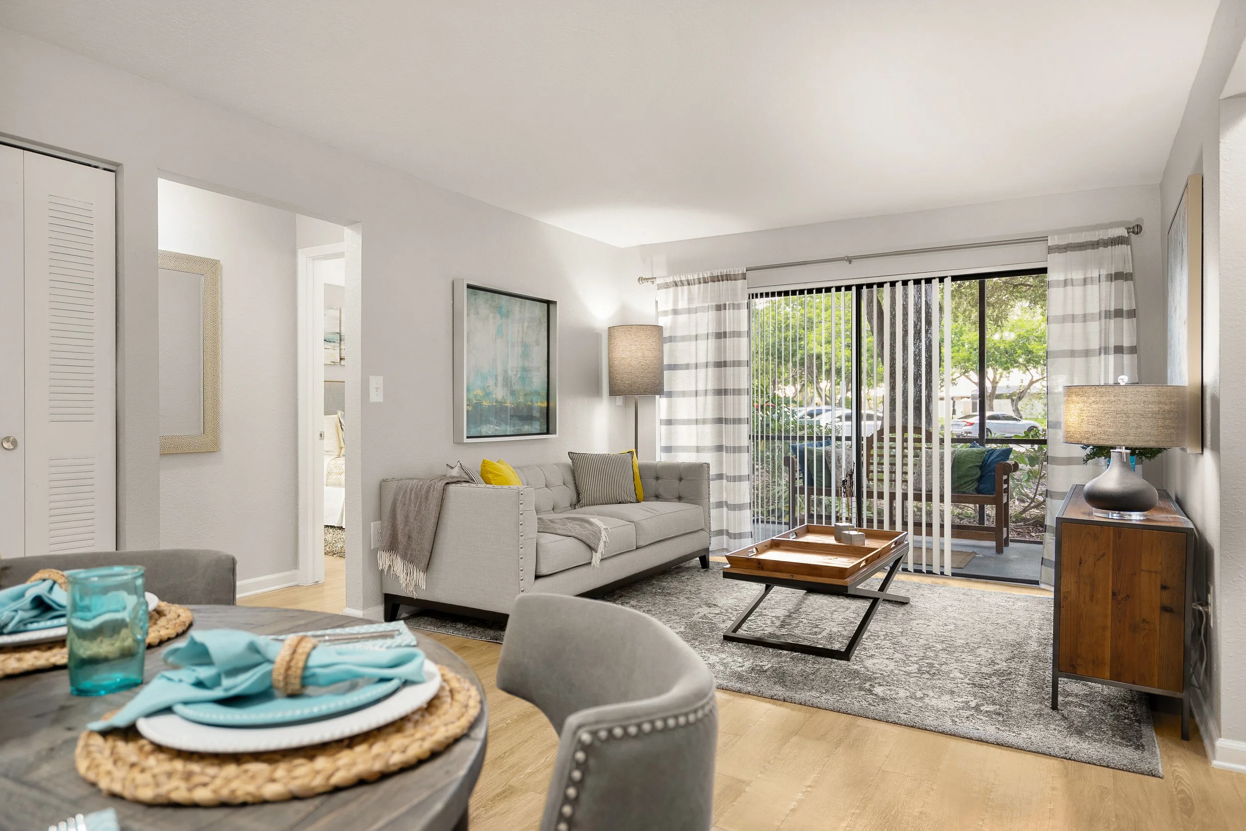 Living room with a beige sofa, wooden coffee table, gray area rug, and sliding glass door with white and gray striped curtains, showing outdoor patio with green trees and parked cars.