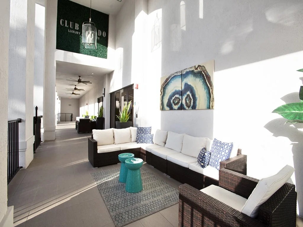 Bright lounge area with white walls, modern wicker furniture including a sofa and chairs with white cushions and blue patterned pillows, two small turquoise side tables, a colorful abstract wall art, and large plants, in a space with natural light an