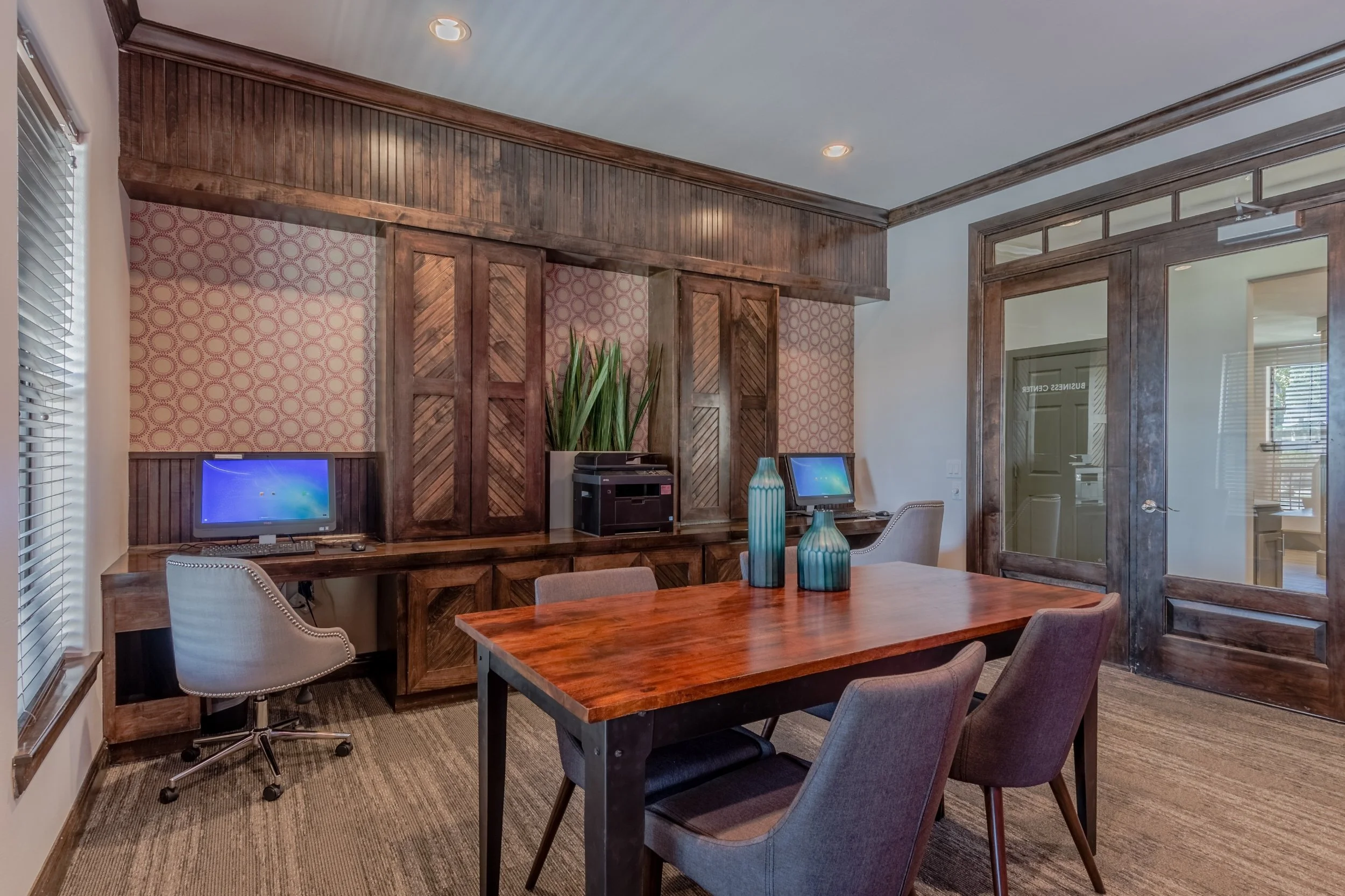 Conference room with wooden accents, a wooden table, four chairs, two computers, decorative vases, and a sliding glass door.