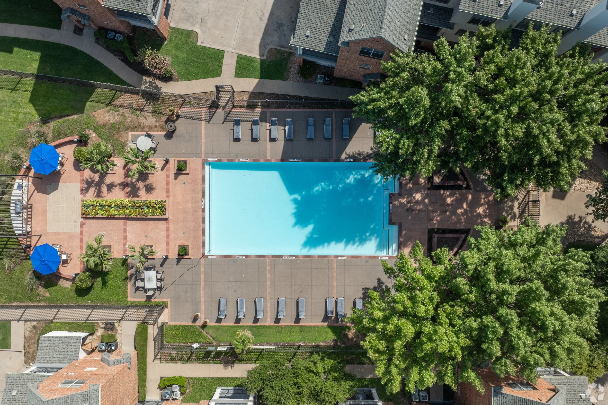 Aerial view of a swimming pool surrounded by a tiled patio, with lounge chairs, umbrellas, trees, and nearby buildings.