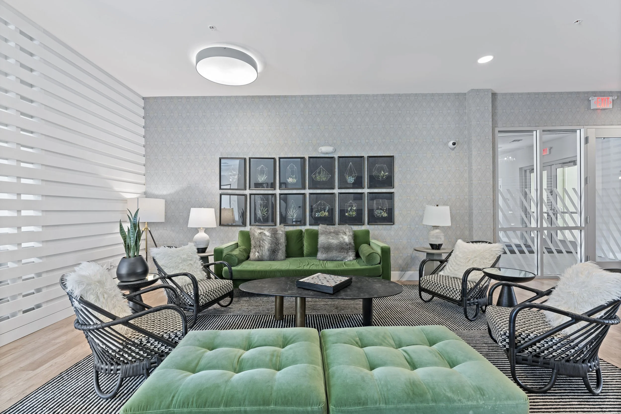Modern lobby with green velvet sofas, black wicker chairs with patterned cushions, white furry pillows, and a black coffee table on a black and white striped rug. Background features a textured gray wall with framed botanical art, three white table l