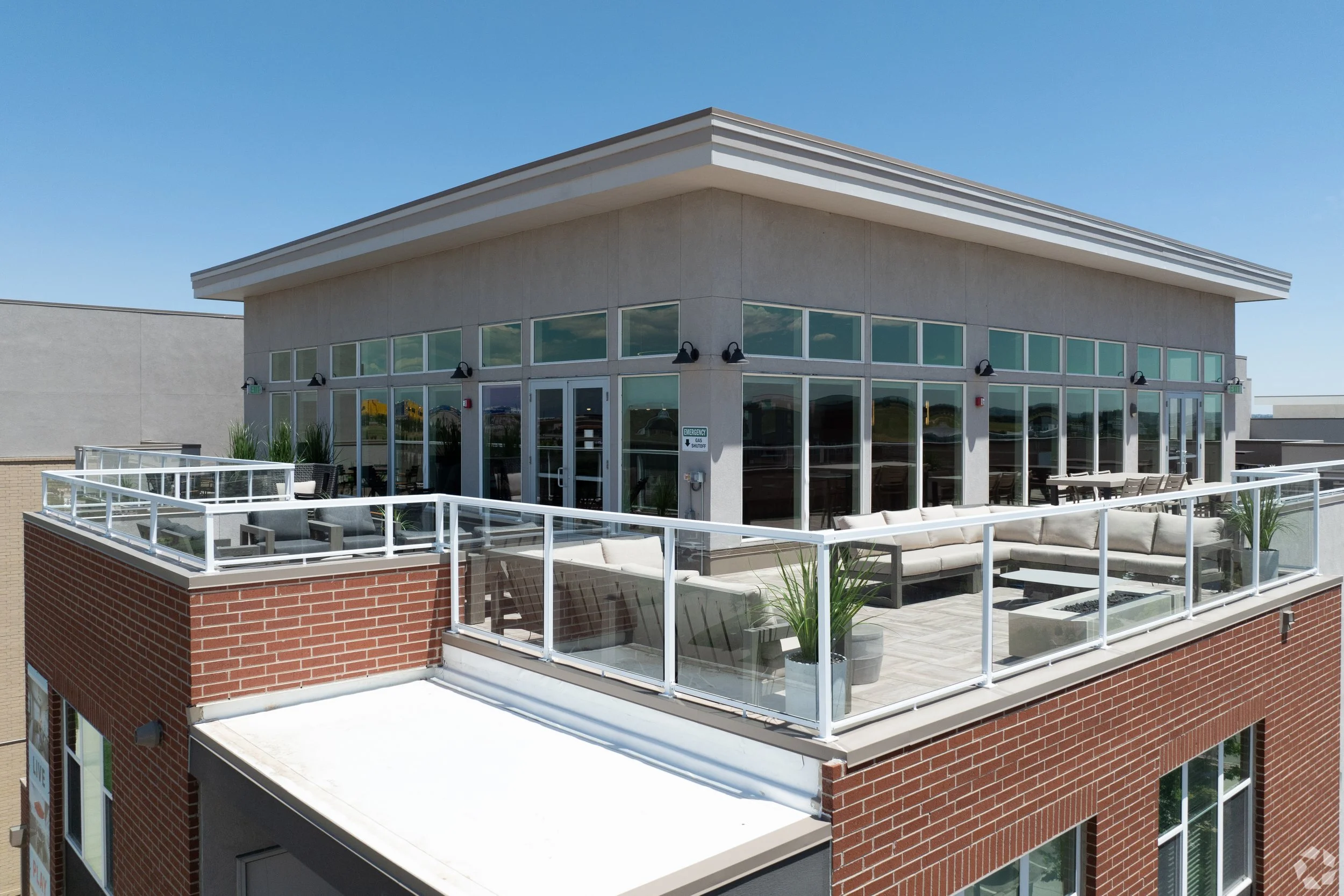 Rooftop patio with glass railing, outdoor seating, and potted plants, atop a brick building under a clear blue sky.
