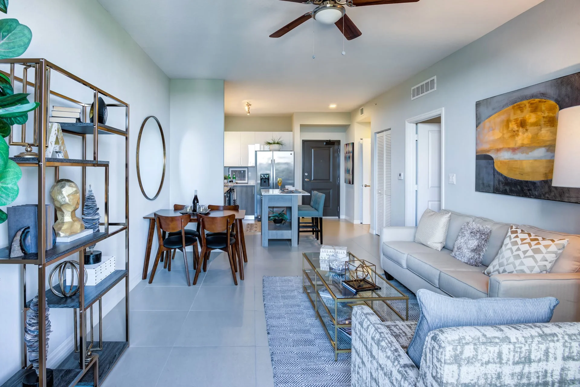 Modern open-plan living room and kitchen with light-colored walls, a ceiling fan, a white sofa with decorative pillows, a glass-top coffee table, a patterned armchair, artwork on the wall, a bookshelf with decor, a dining table with chairs, and a kit