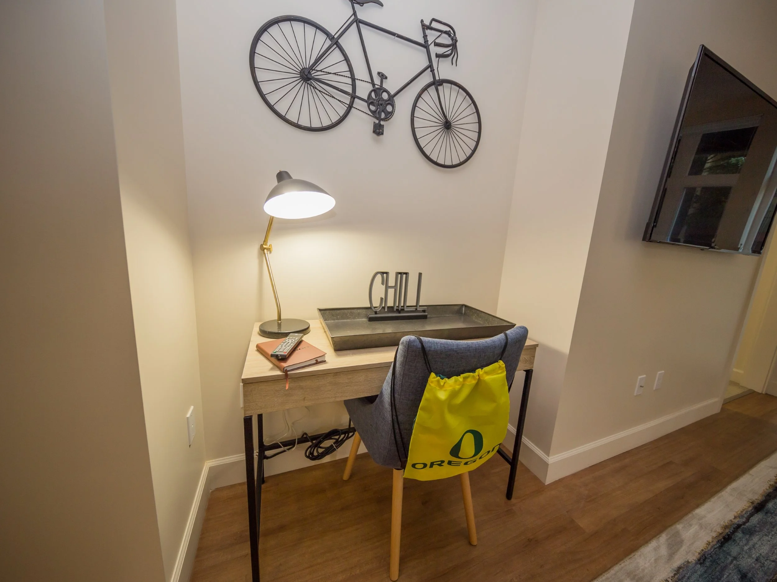 A small desk with a gray chair, a yellow Oregon bag hanging on the back of the chair, a desk lamp, a remote, a notebook, and a tray with a decorative word "CHILL" on it. There is a wall-mounted black bicycle sculpture and a wall-mounted television in