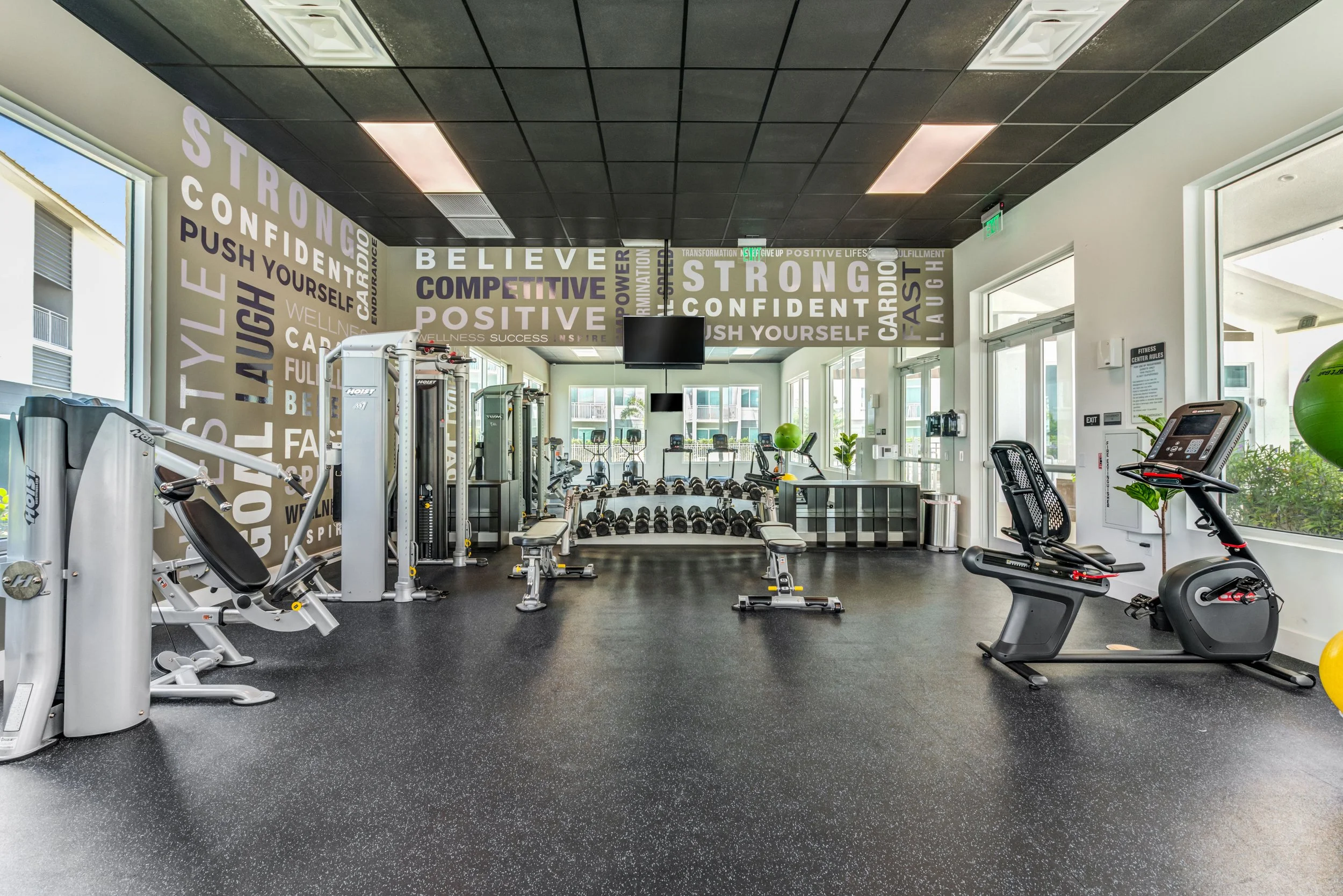 Modern gym with workout equipment including stationary bike, weight machines, dumbbells, and benches, with motivational words on the wall and large windows providing natural light.
