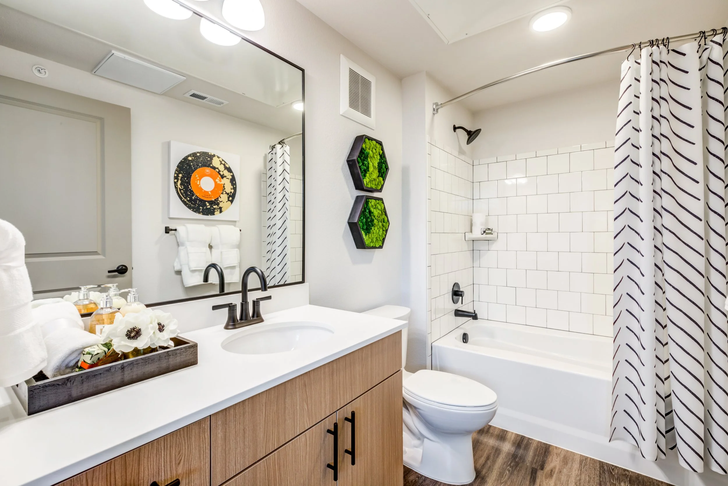 Modern bathroom with white vanity, large mirror, white subway tile shower, wooden flooring, and decorative wall art.