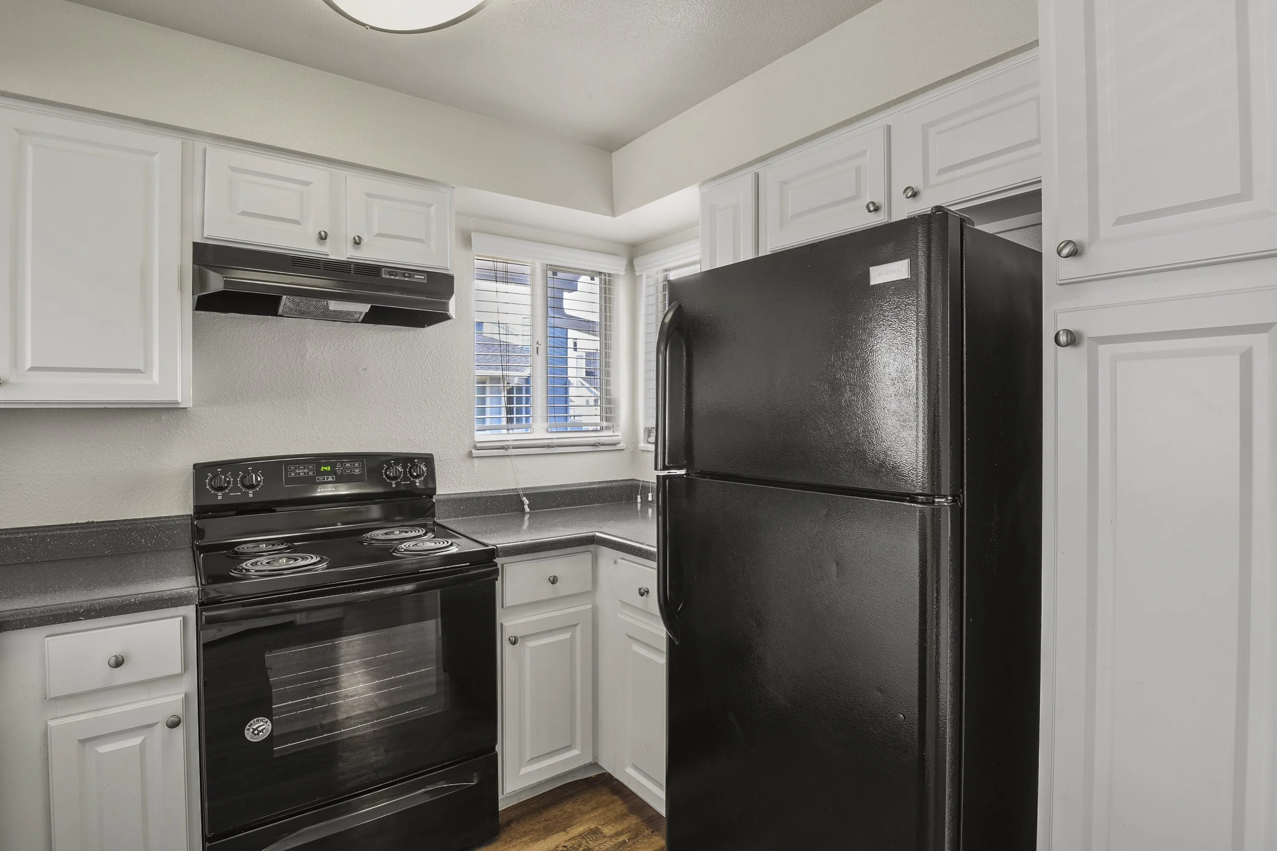 A small kitchen with white cabinets, a black refrigerator, a black stove, and a window with white blinds.