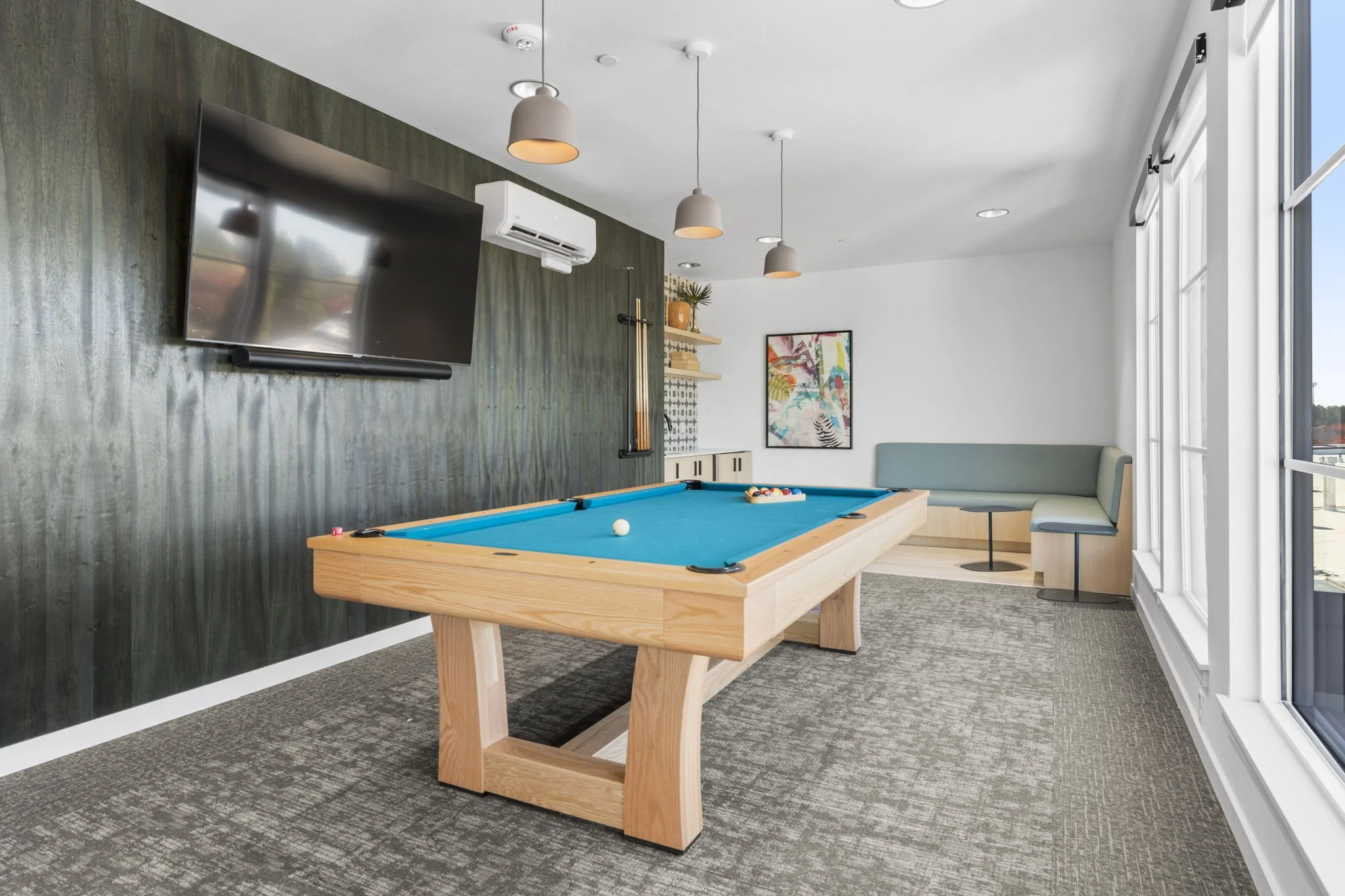 Modern recreational room with a blue pool table, wall-mounted TV, large windows, colorful art, and seating area.