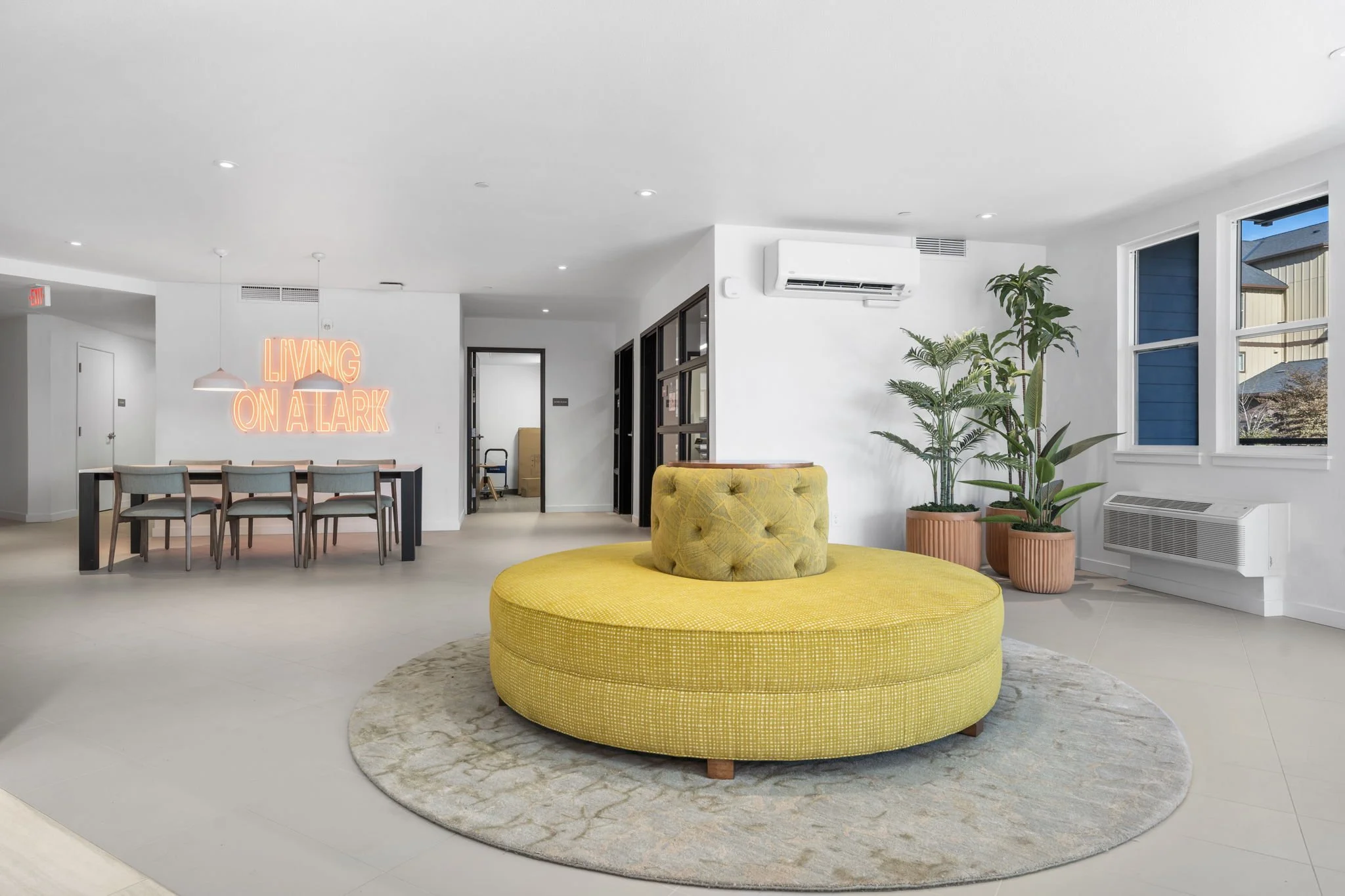 Modern living room with yellow circular sofa on a patterned rug, potted plants, large windows with blue shutters, and a neon sign that says 'Living on Clark' in the background.