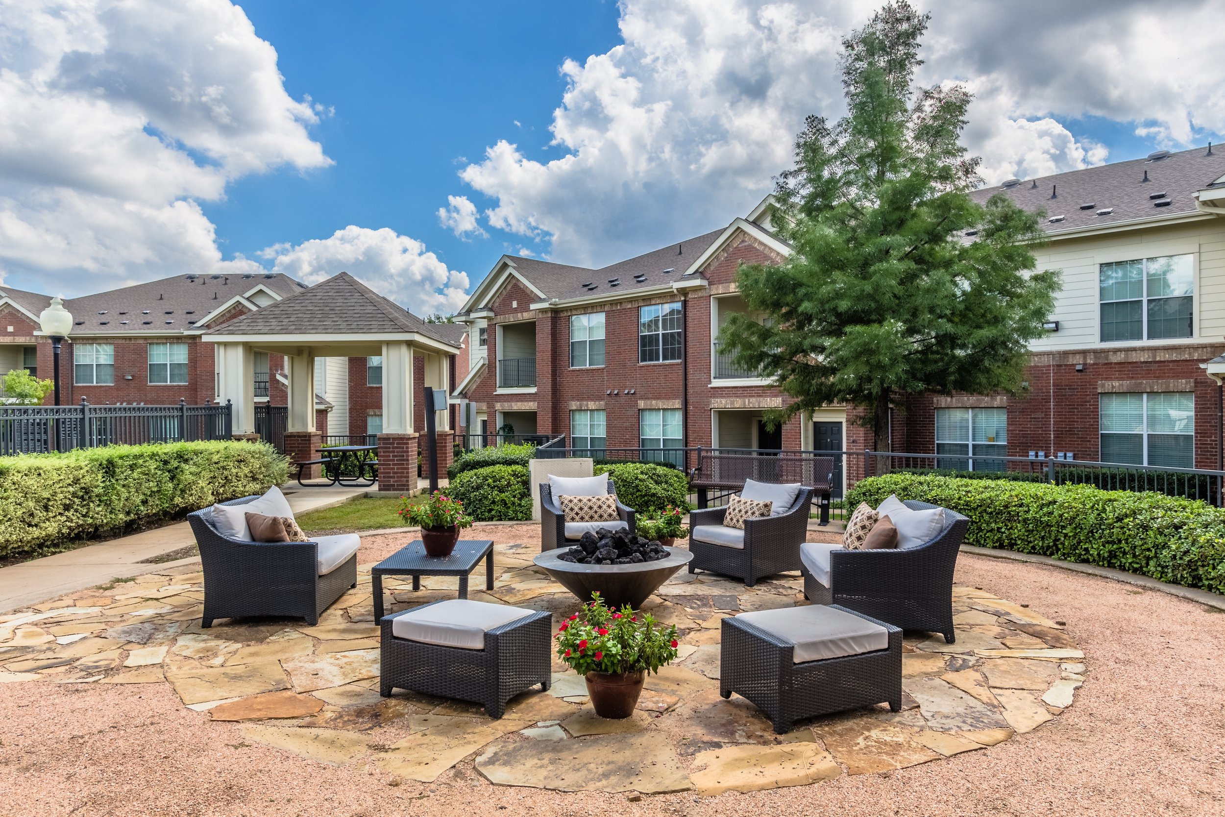 A residential apartment courtyard with circular arrangement of outdoor chairs, potted plants, a fire pit, surrounding hedges, and brick buildings with balconies under a partly cloudy sky.