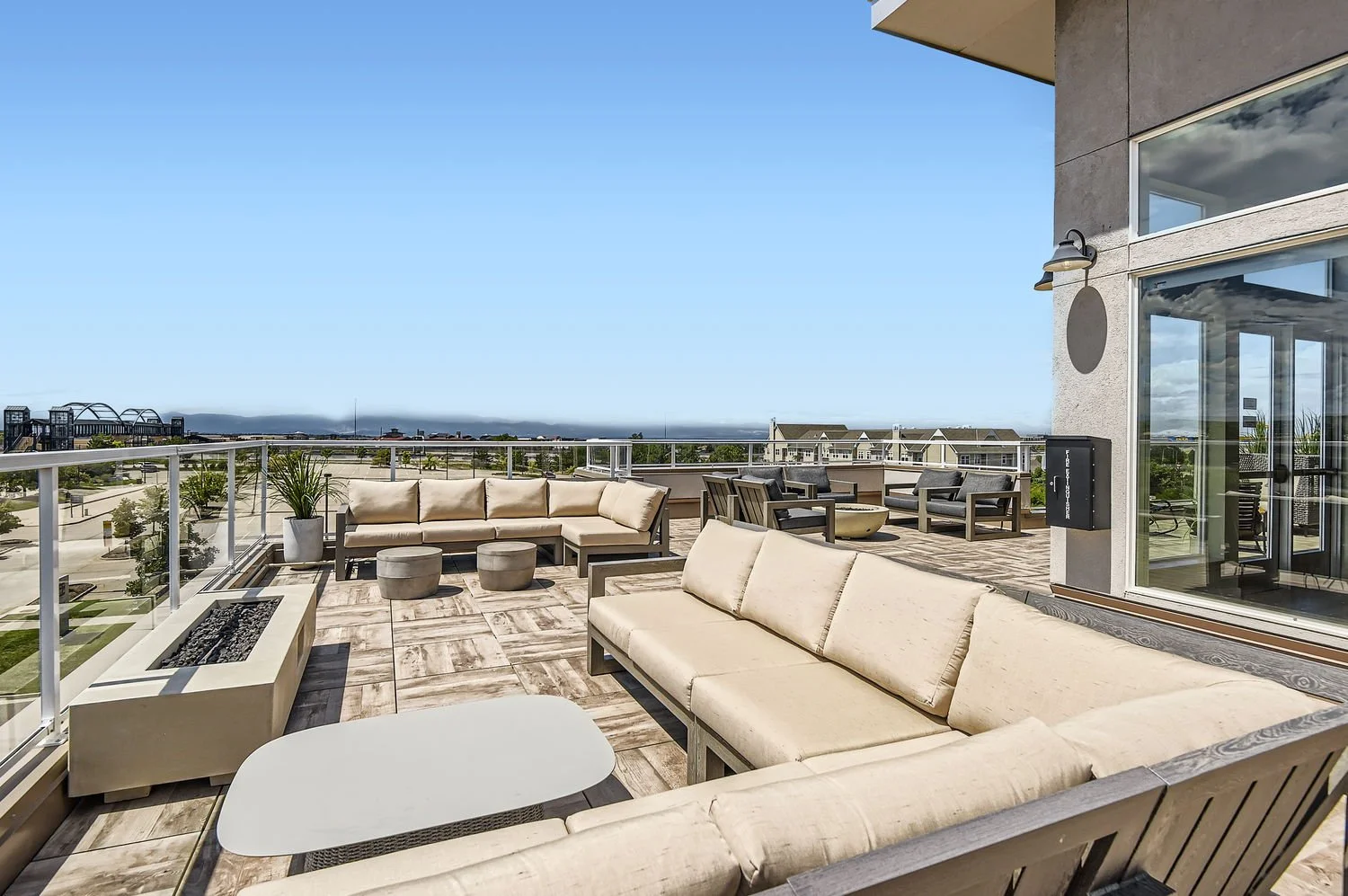 Rooftop terrace with beige outdoor sofas, black armchairs, potted plants, and a fire pit, overlooking a cityscape with a clear blue sky.
