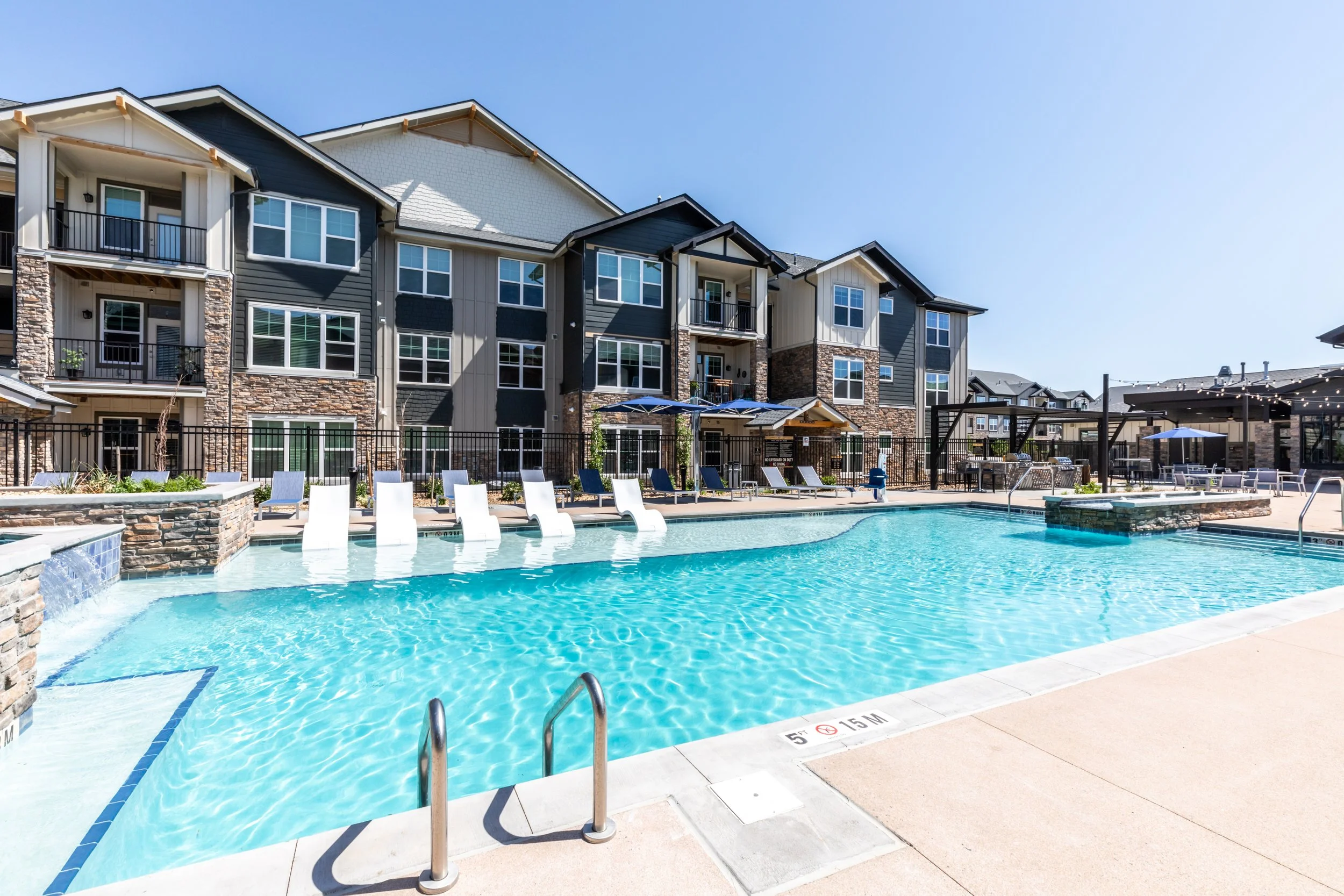 Outdoor swimming pool at an apartment complex with lounge chairs, tables with umbrellas, and multi-story residential buildings in the background.