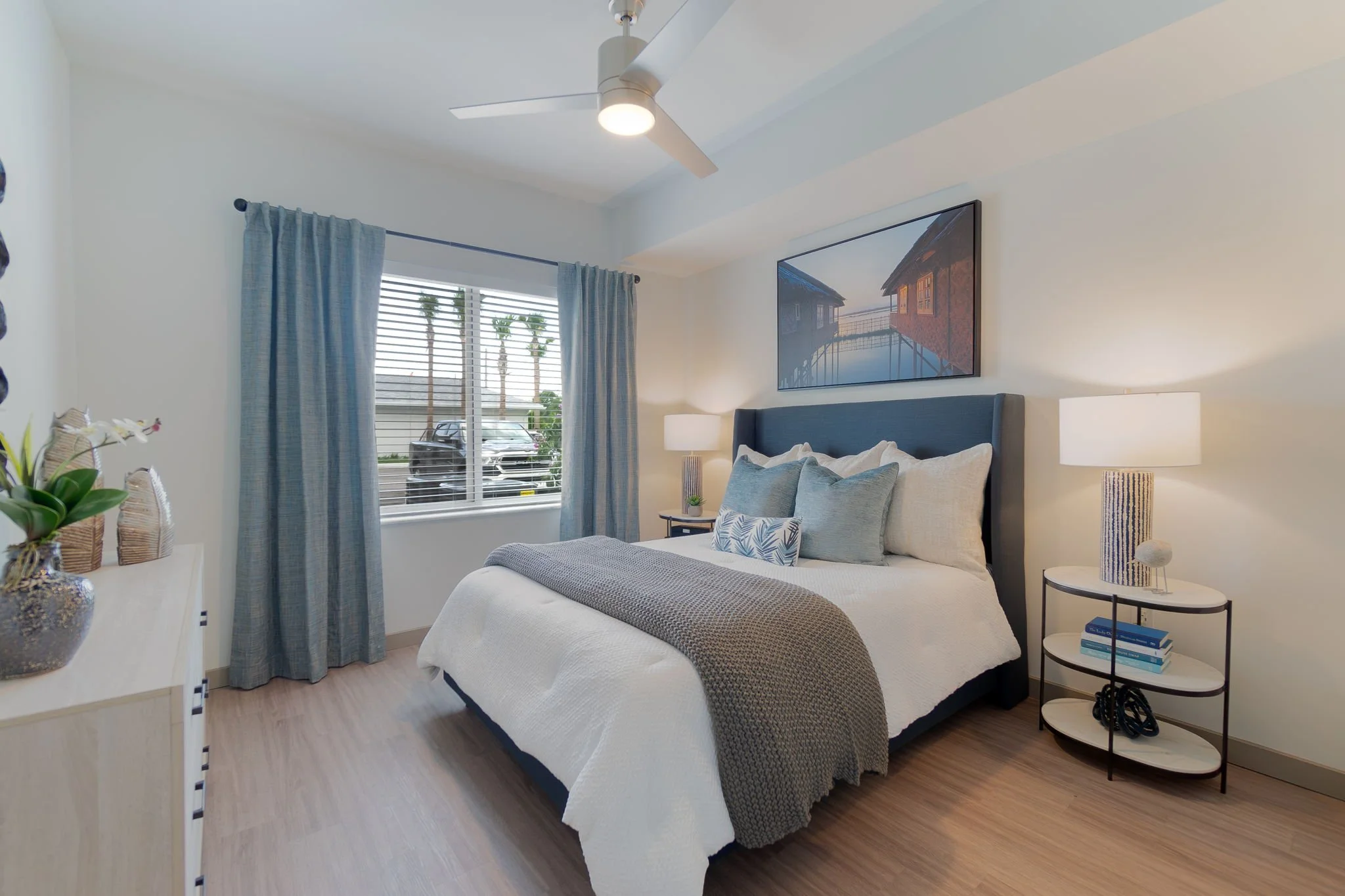 Modern bedroom with a bed, blue headboard, white bedding, and gray blanket. Nightstand with a lamp, books, and decorative items. Window with blue curtains showing cars and palm trees outside. Wall art above the bed depicts waterfront houses.