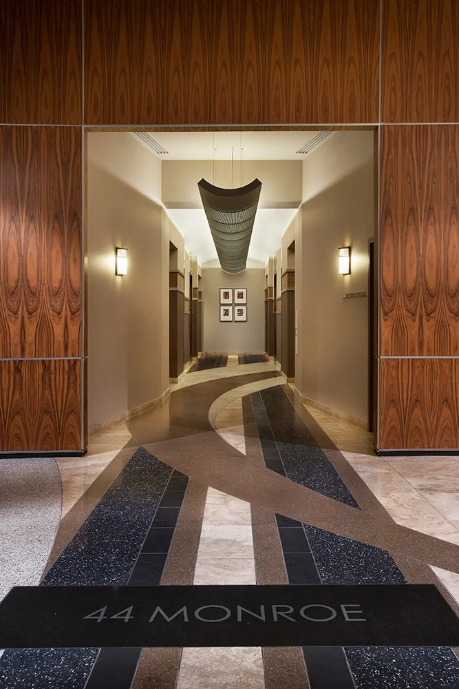 Hotel hallway entrance with a black doormat reading '44 Monroe', wood-paneled walls, and patterned flooring leading to framed artwork on the back wall, with modern light fixtures along the sides.