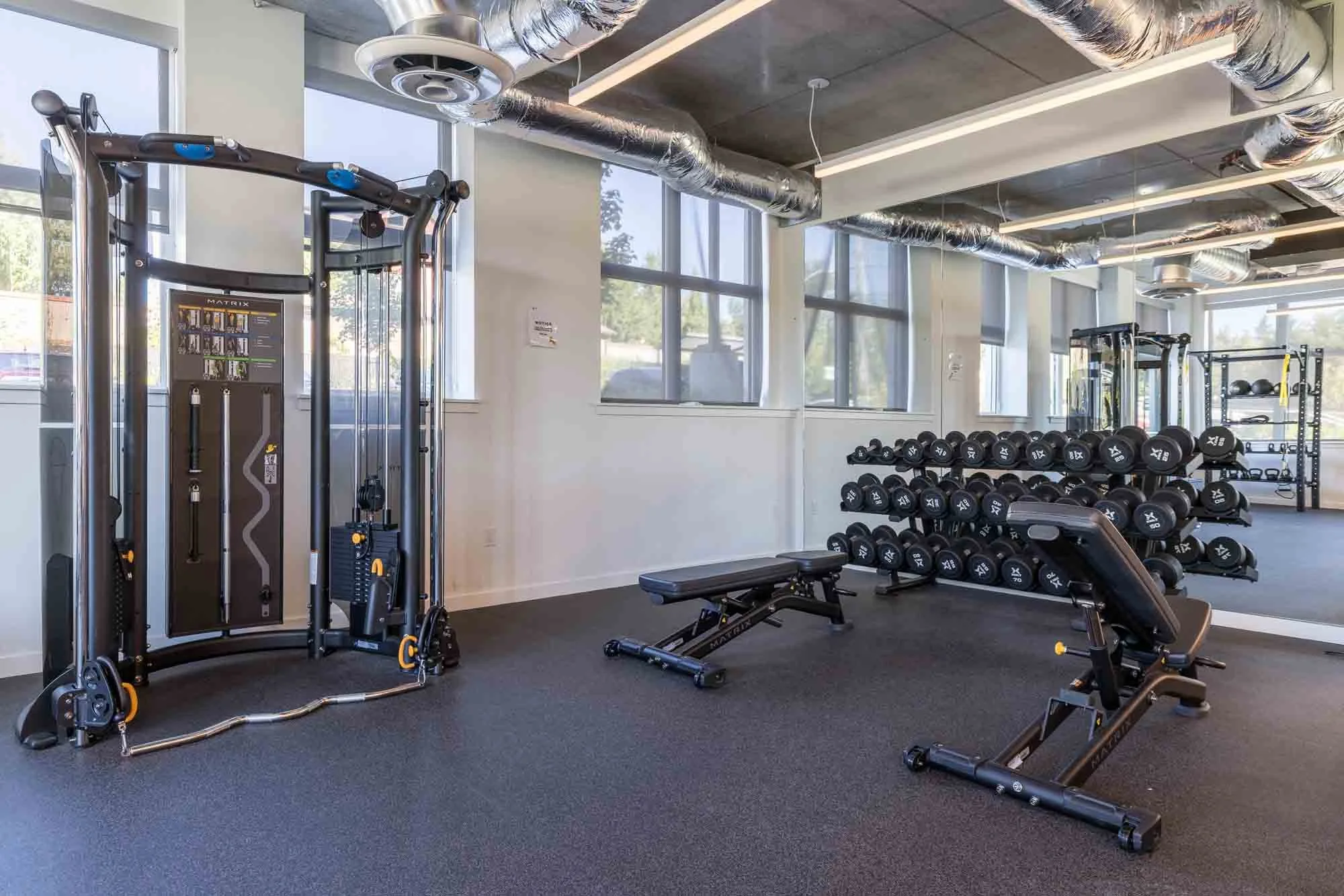 A modern gym with various exercise equipment including a cable machine, dumbbells on racks, a workout bench, and a workout chair, with large windows and visible ductwork on the ceiling.