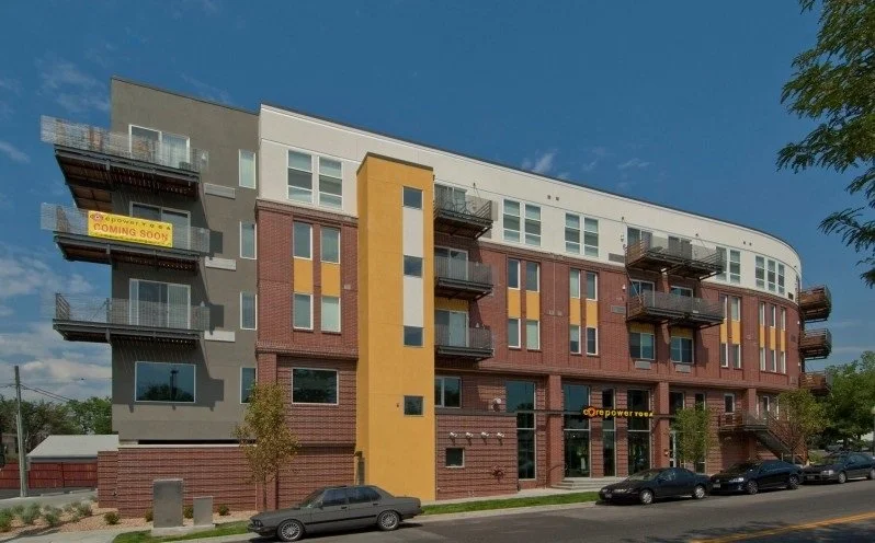 Modern multi-story apartment building with balconies and a sign that reads 'Core Power 102' on the front, on a city street with parked cars and trees.
