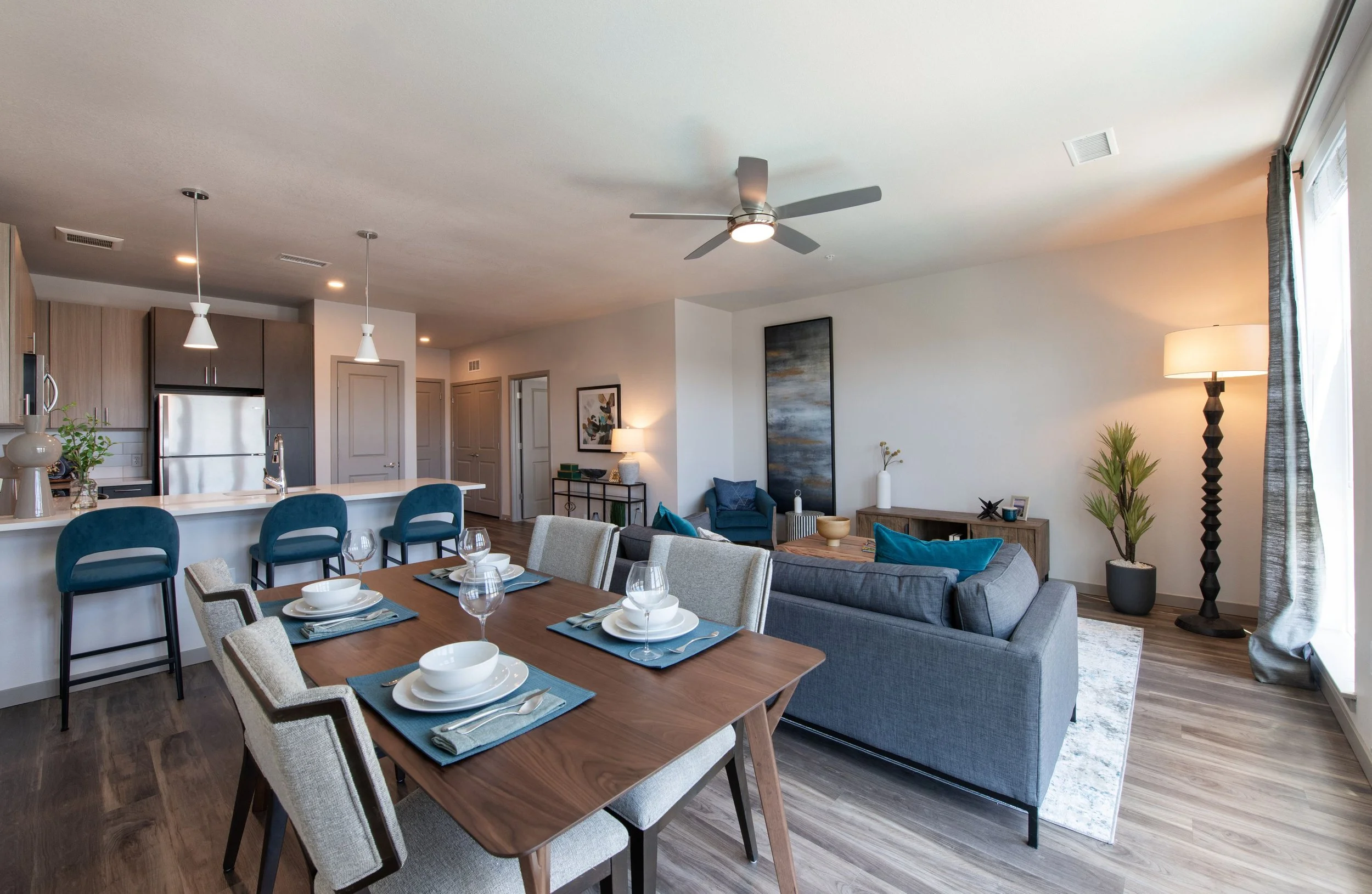 Modern open-concept living and dining area with wood flooring, a gray sofa, a wooden dining table set for four, a kitchen with a white countertop, blue barstools, and decorative plants and artwork.