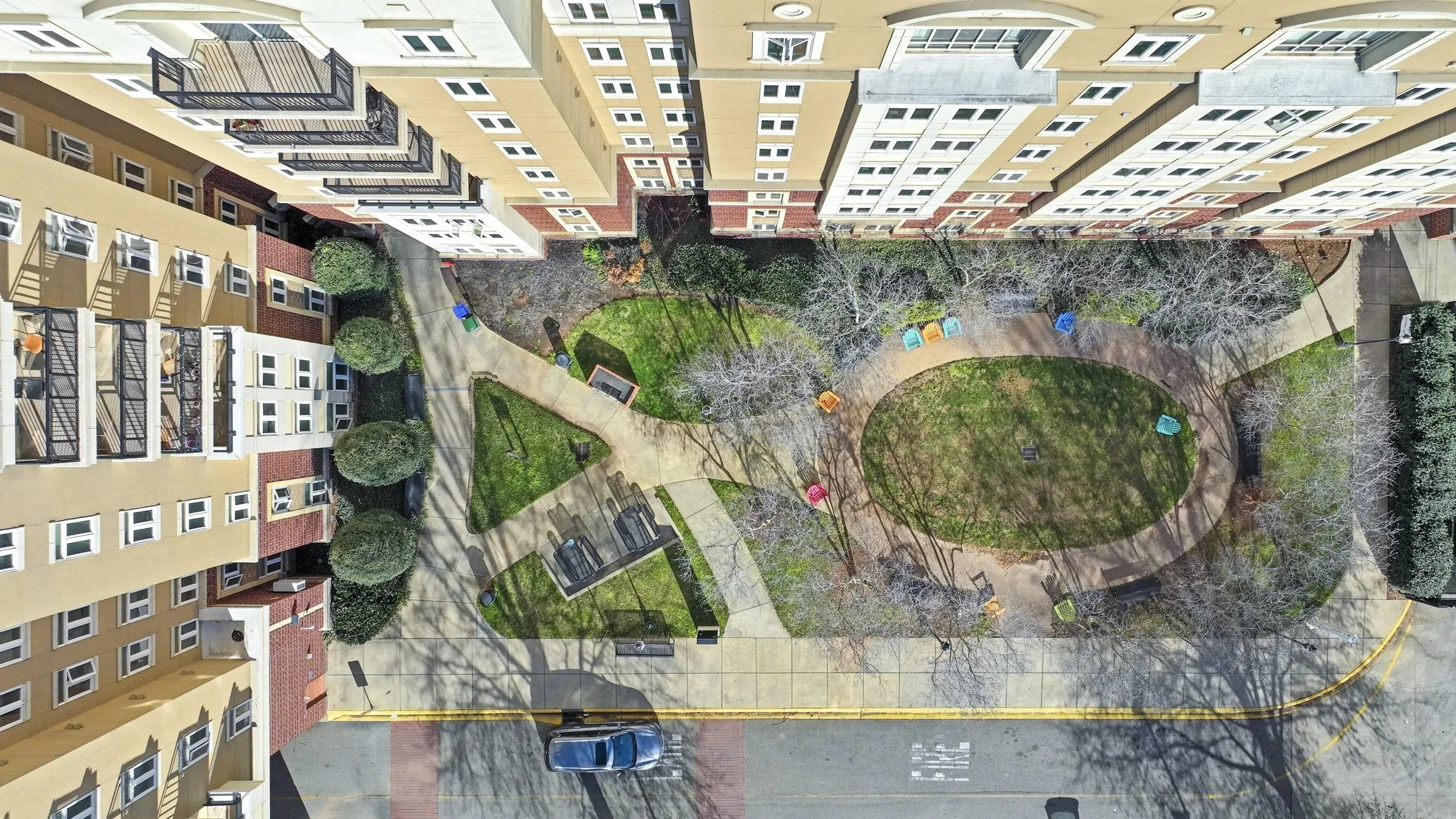 An aerial view of a courtyard between apartment buildings with a walking path, grassy areas, benches, and leafless trees.