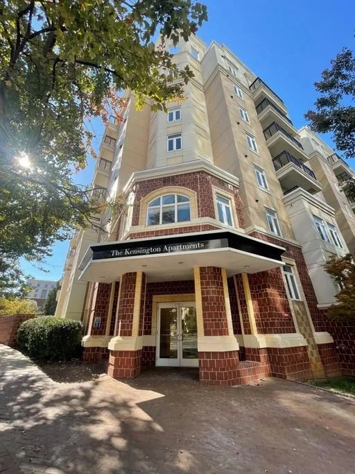 Exterior view of a tall residential building called The Kensington Apartments, with brick and beige siding, balconies, and a clear blue sky.