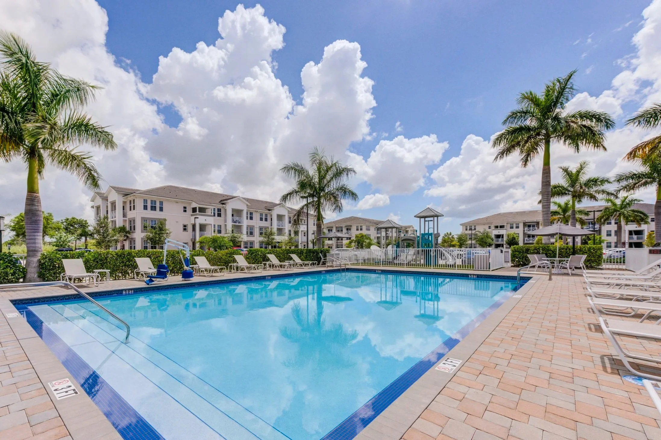Luxury apartment complex outdoor swimming pool with palm trees, lounge chairs, umbrellas, and a children’s playground under a partly cloudy sky.