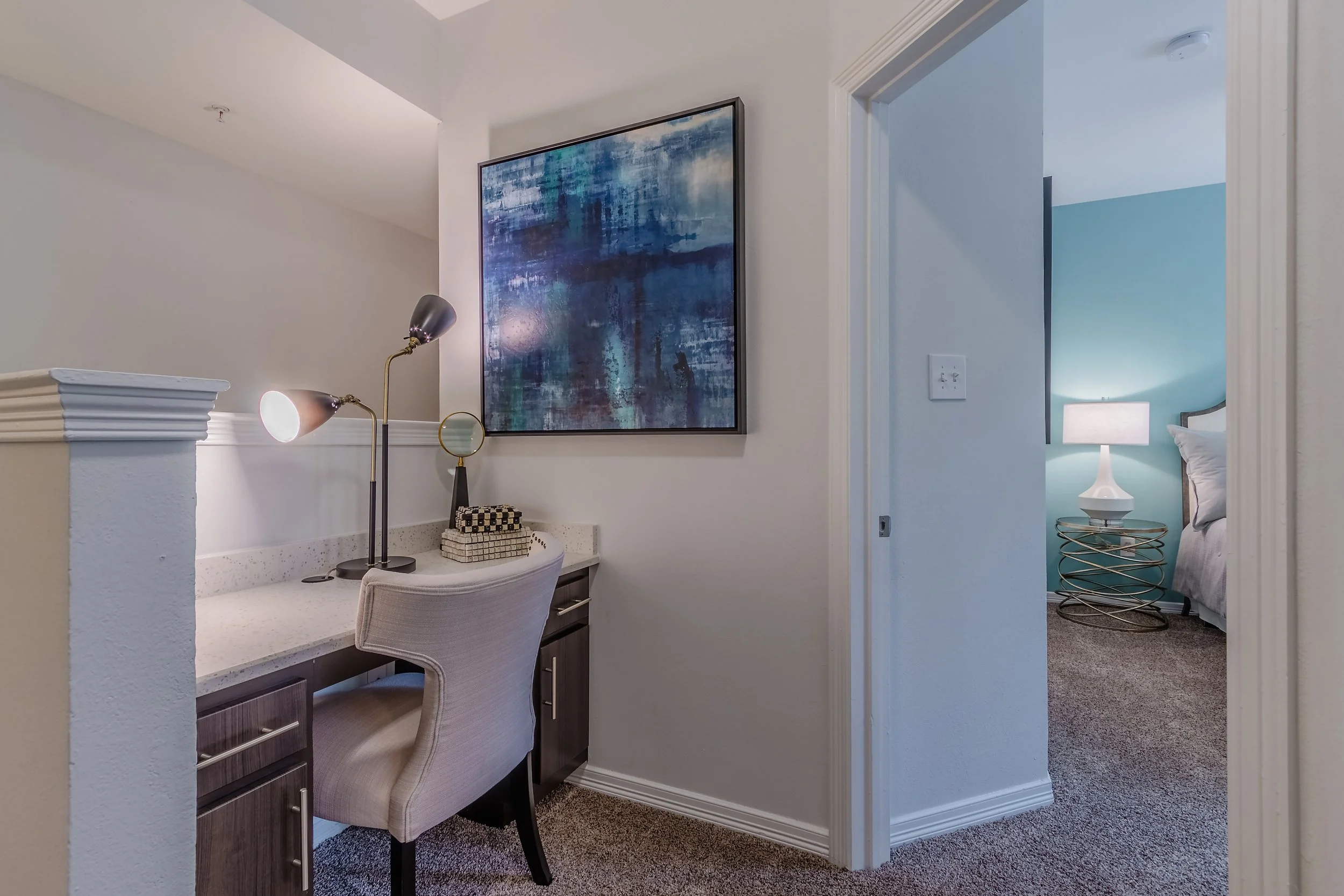 A home office with a desk, beige chair, modern desk lamp, abstract wall art, and a view into a bedroom with a glowing lamp on a side table and a bed with white linens.