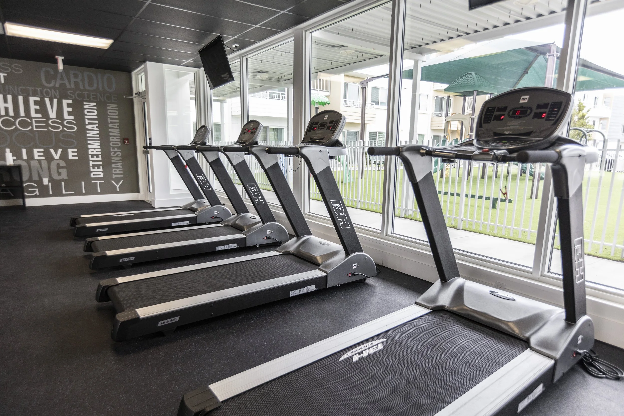 Five treadmills positioned in a row in a gym with large windows revealing a fenced outdoor area.