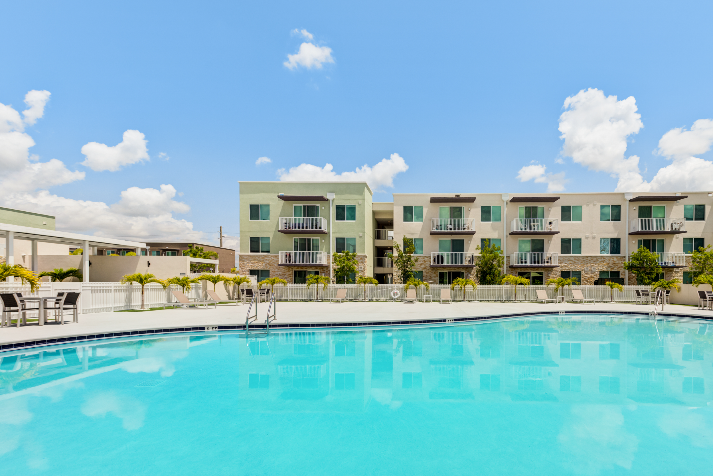 An outdoor swimming pool in front of a modern apartment complex with green and beige exteriors, surrounded by lounge chairs and palm trees under a blue sky with scattered clouds.