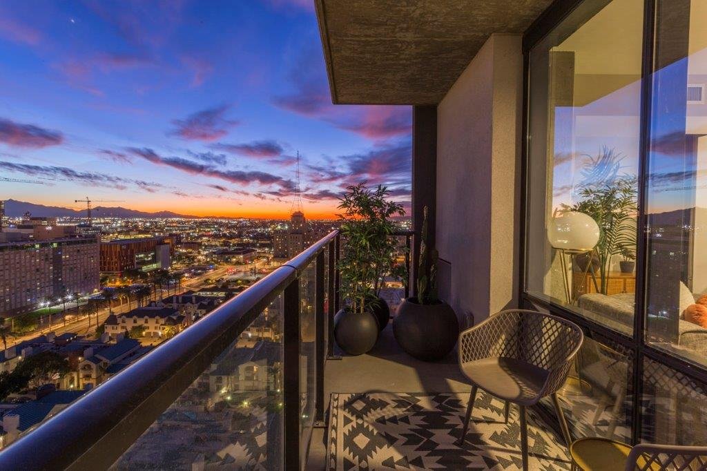 Balcony view of a city skyline during sunset, with skyscrapers and clouds, balcony furniture, potted plants, and a room visible through a glass door.