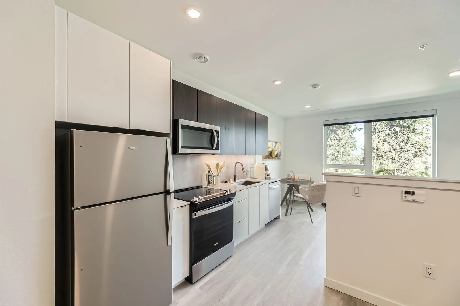 Modern kitchen with stainless steel refrigerator, microwave, stove, and white cabinets, adjacent to a dining area with a window overlooking trees.