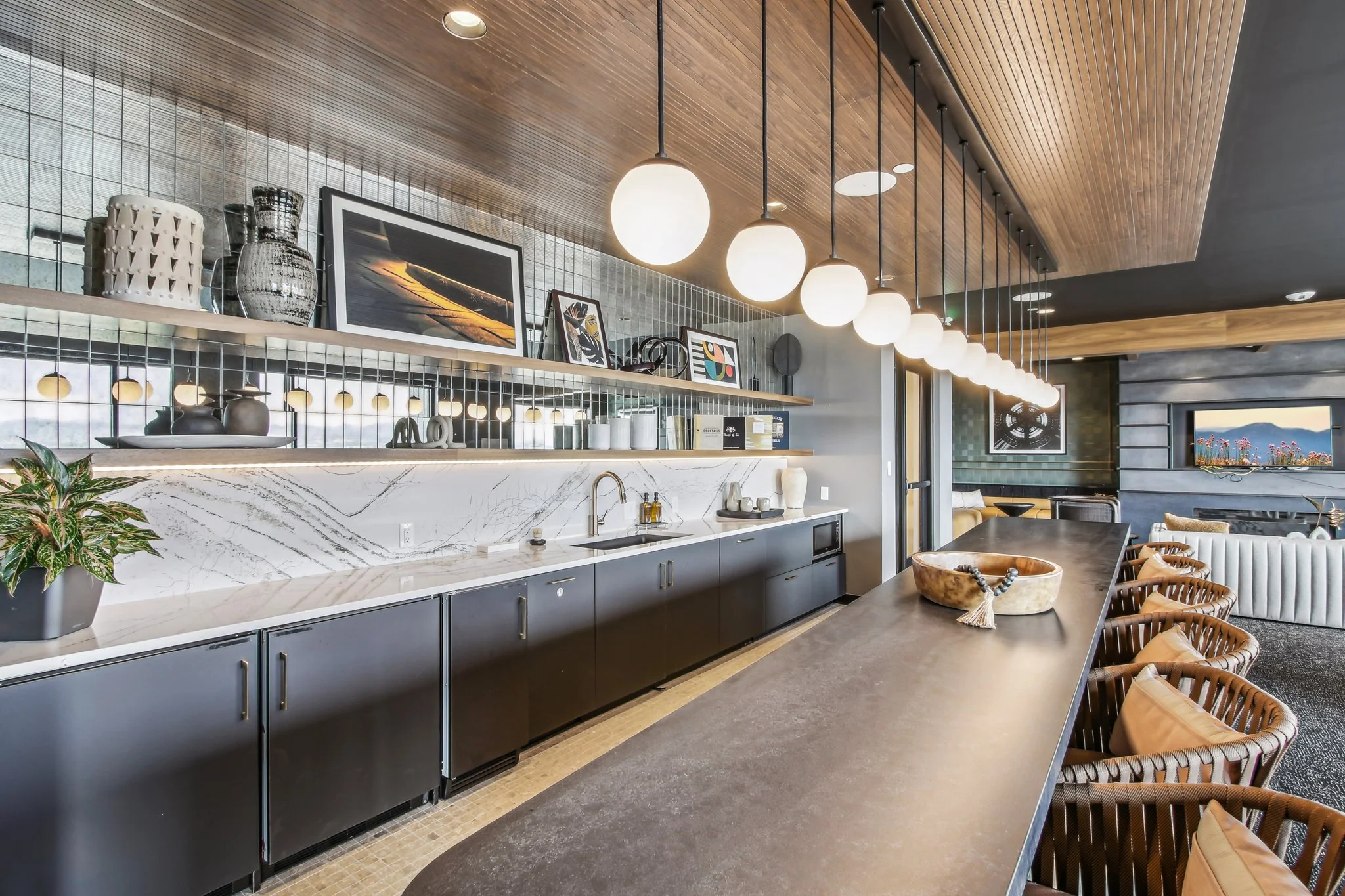 Modern kitchen with black lower cabinets, a white marble countertop with gray veining, an open shelf with framed photos, vases, and decorative objects, white globed pendant lights hanging above a long dark counter with woven chairs, a plant on the co