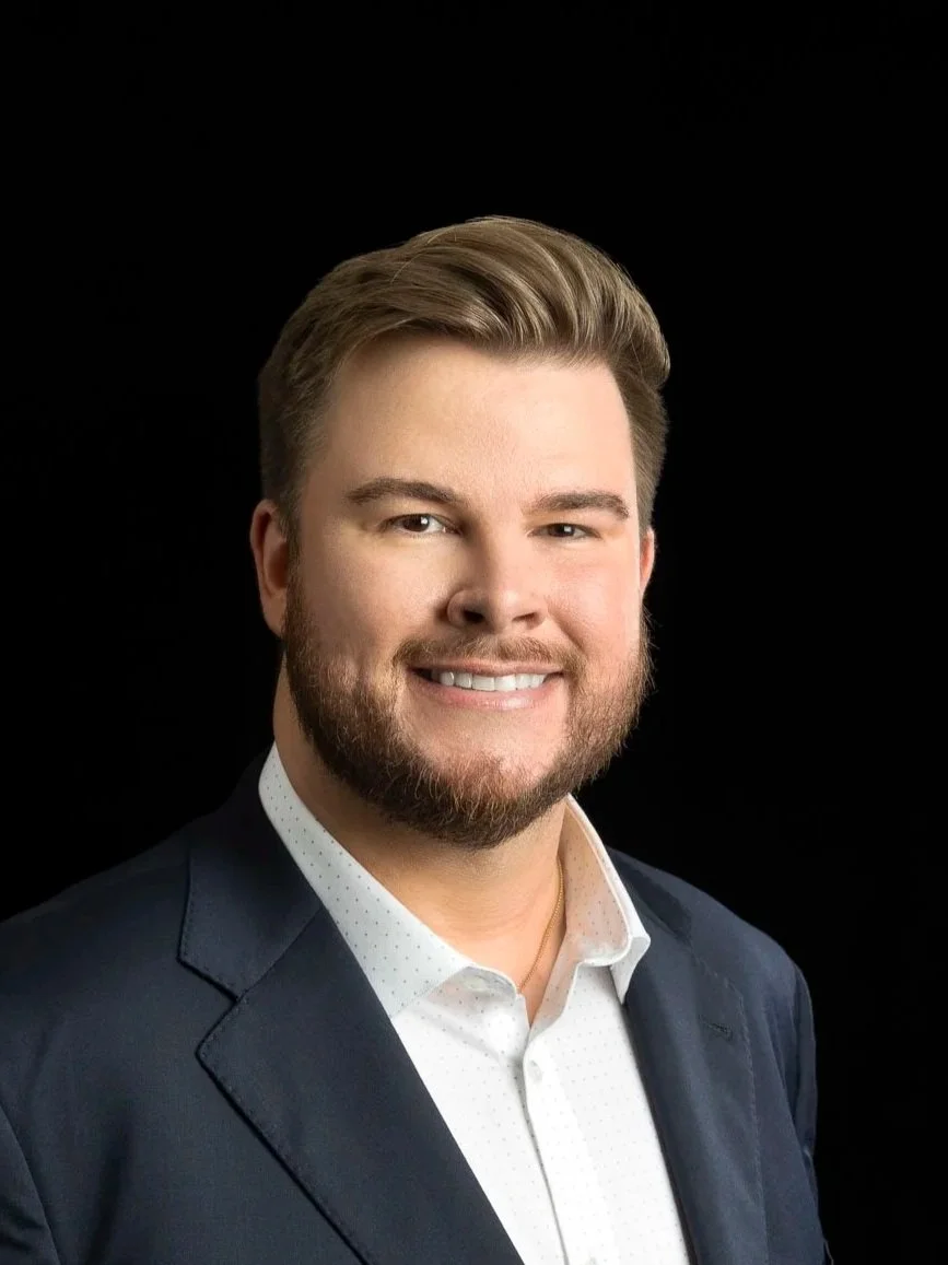 Headshot of a smiling man with light brown hair and beard, wearing a navy blazer and white shirt against a black background.