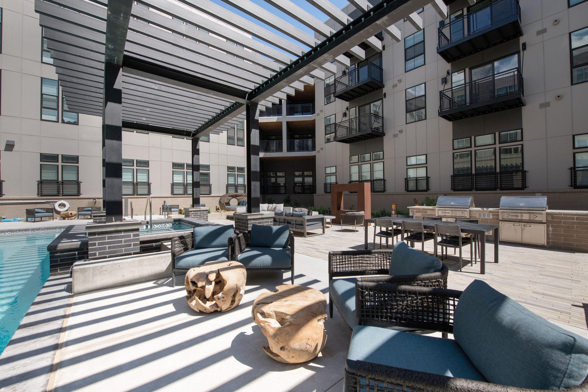 Modern apartment complex rooftop with pool, outdoor seating, and barbecue grills, shaded by a pergola with shadows cast on the deck.