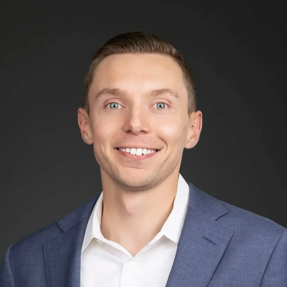A professional portrait of a young man with blonde hair, blue eyes, wearing a navy suit and white shirt, smiling against a dark background.