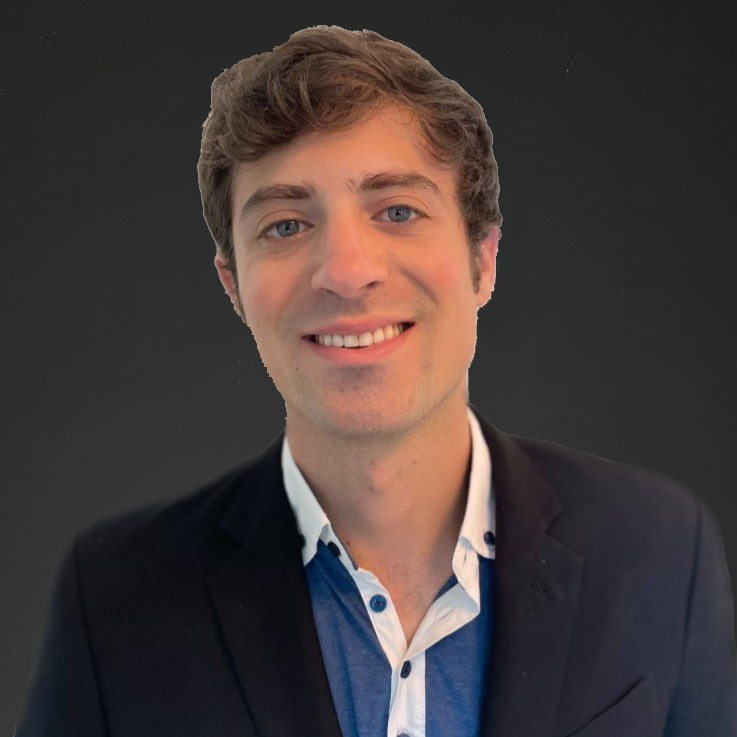 Portrait of a young man with light brown hair, blue eyes, wearing a dark blazer over a shirt, smiling against a dark background.