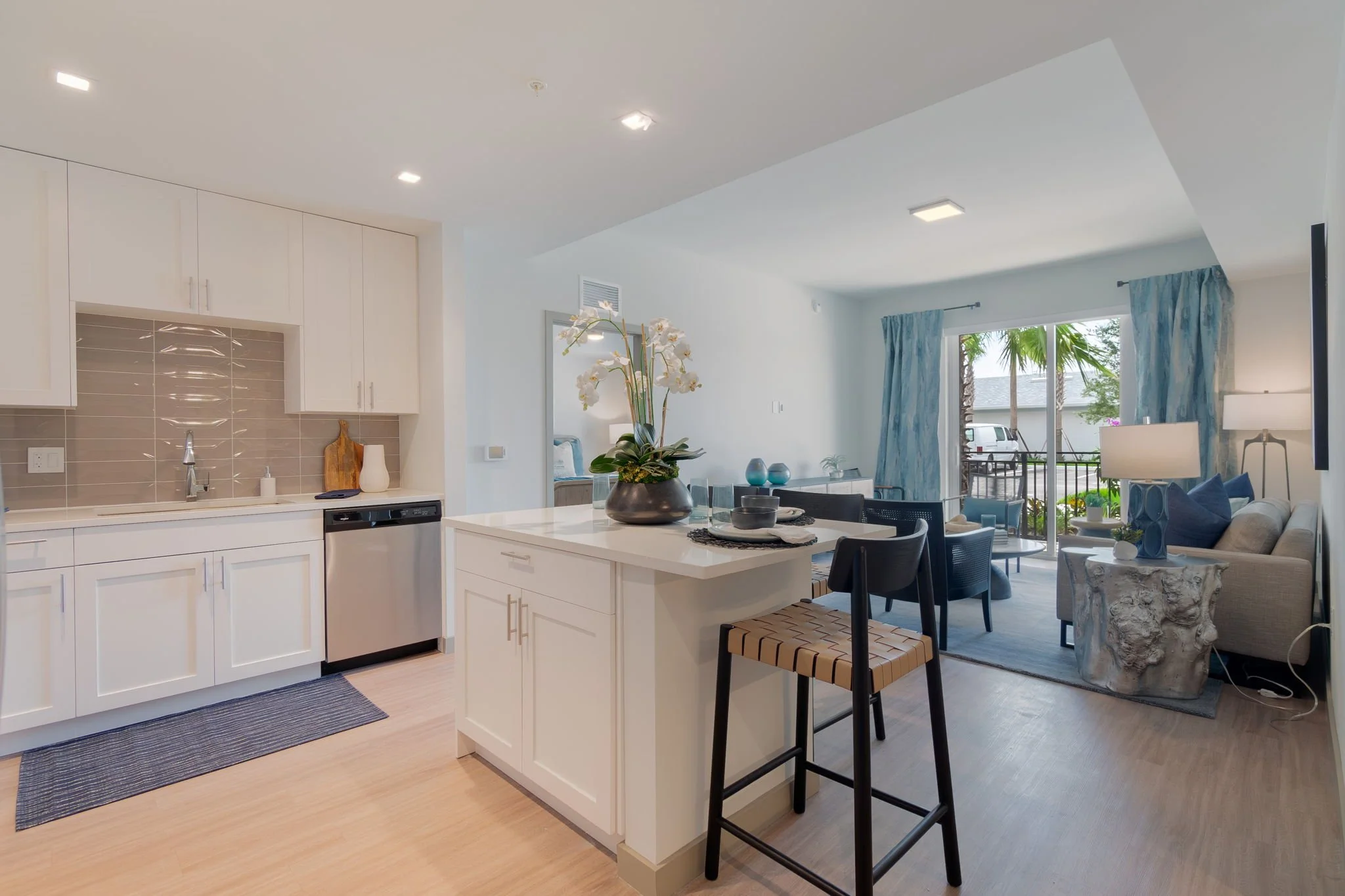 Modern open-concept living space with a kitchen and sitting area. The kitchen has white cabinets, a gray backsplash, and a small island with a decorative flower arrangement. The sitting area includes a beige couch with blue pillows, a stone side tabl